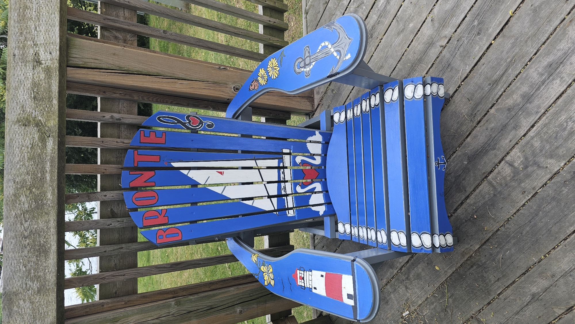 This media file shows a close-up, eye-level, top-down shot of a brightly painted Adirondack chair placed on a wooden deck. The chair is predominantly blue and grey with various nautical-themed designs painted on it. On the backrest, there is a grey anchor with a white rope against a blue background, surrounded by a pattern of white circles with grey outlines. On the seat, there is a large white sail on a boat, with two red hearts depicted as swans. The text "BRONTE" is painted in red capital letters on the slats of the seat. The armrests also feature painted designs. The left armrest has a yellow flower and a white butterfly on a blue background, while the right armrest displays a red and white lighthouse against a blue background.
The chair is positioned on a weathered wooden deck, with glimpses of green grass and trees in the background, suggesting an outdoor setting. The lighting indicates it is daytime, likely sunny given the clear visibility. The overall impression is of a charming, personalized piece of outdoor furniture, likely located in a coastal or waterfront area given the nautical motifs. The presence of "BRONTE" suggests a connection to the Bronte Creek Provincial Park or the Bronte area of Oakville, Canada, a location known for its waterfront. There is no discernible activity or event taking place, as the chair is unoccupied.