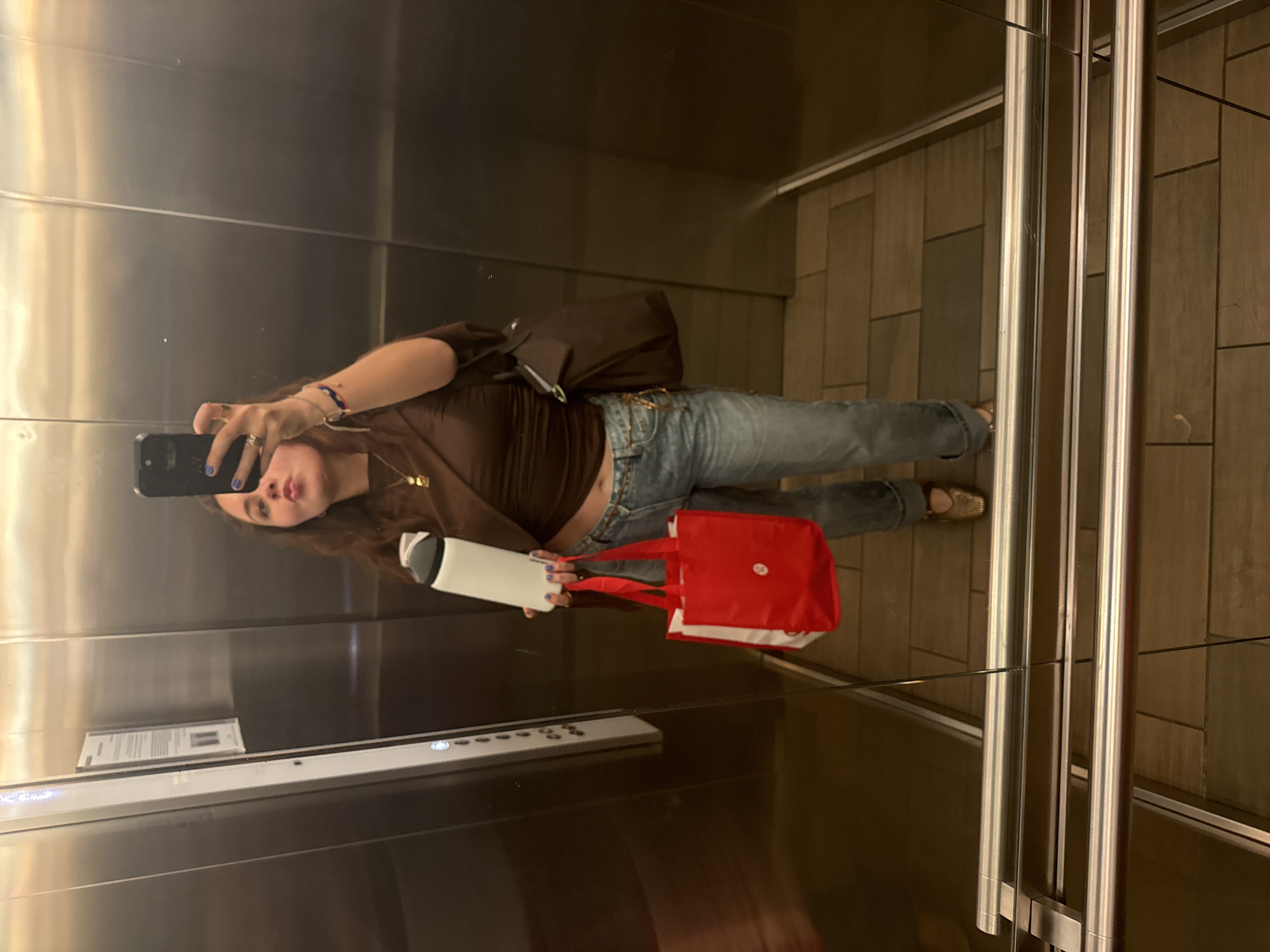 The image is a selfie taken in what appears to be the interior of an elevator, possibly in Mexico City, Mexico, but lacking contextual clues to confirm. The photographer is a young woman holding a cell phone, presumed for taking the picture. She has brown hair and is wearing a brown top, blue jeans, and is carrying a red and white tote bag and a white water bottle. The elevator's interior walls consist of reflective metal paneling, showing the woman's reflection and a control panel. The floor of the elevator is covered with brown tiles. The elevator has metal bars near the door and a notice or sign posted on the interior wall.