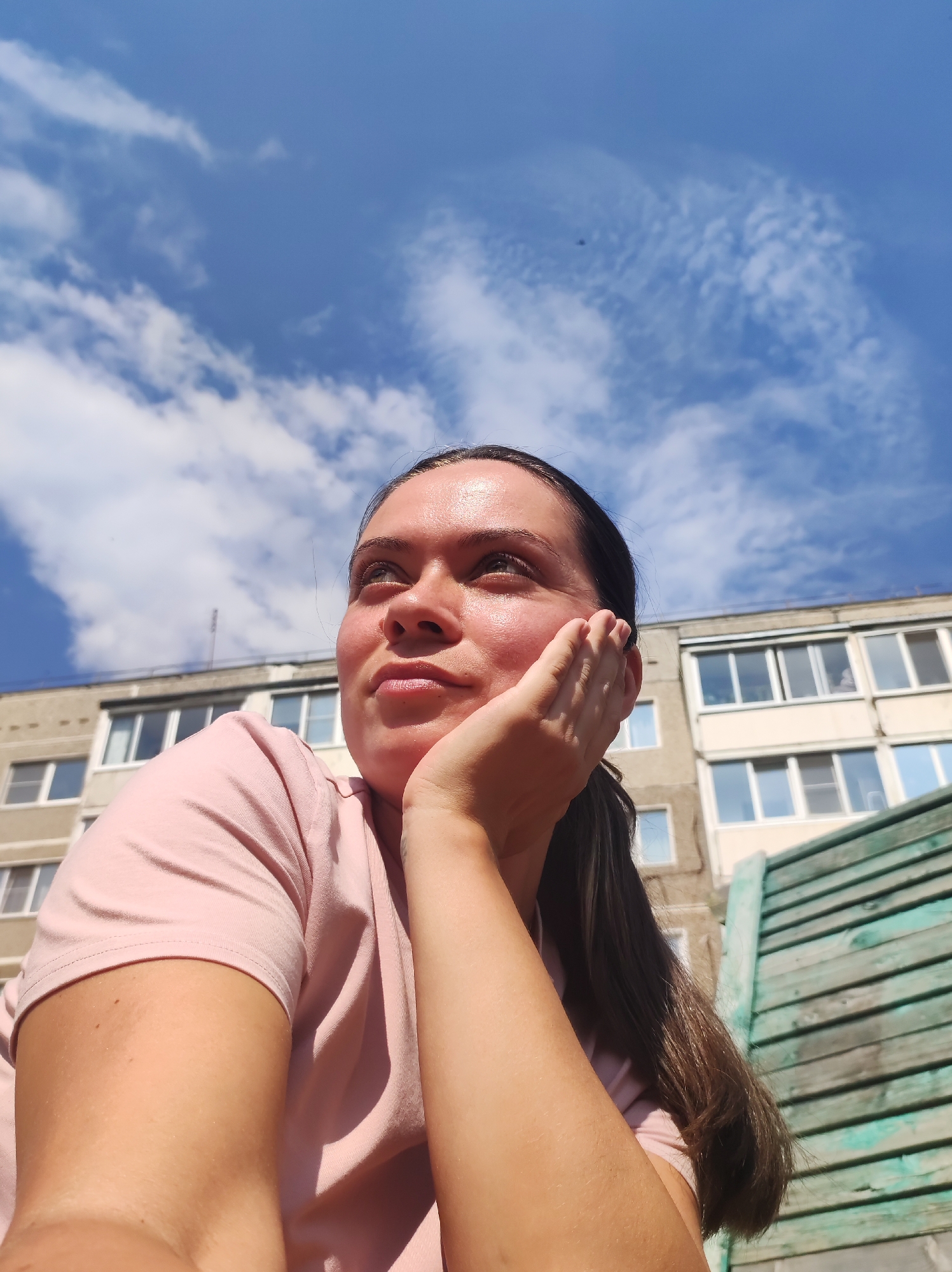 A young woman with dark hair and her hand on her cheek looks upwards against a bright blue sky with wispy clouds. She is wearing a pink t-shirt. Behind her, a multi-story building with many windows is visible. To the right, a weathered green wooden fence is in the foreground. The scene appears to be outdoors, likely in an urban environment in Chaikovsky, Russia, given the architectural style of the building.
