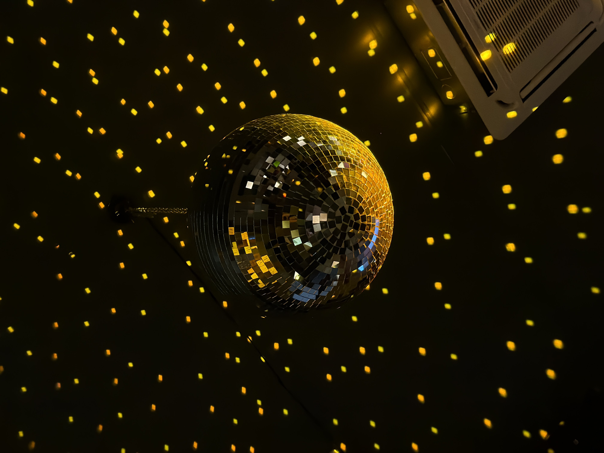 A disco ball hangs from the ceiling, reflecting light in small squares onto the dark walls and the ceiling-mounted air conditioning unit. The air conditioning unit is white. The disco ball is gold and silver. There are visible power cords. The location of the image is not deducible from the given information. Therefore, the location in the city of Krasnoyarsk, Russia, is not discernible from the image.