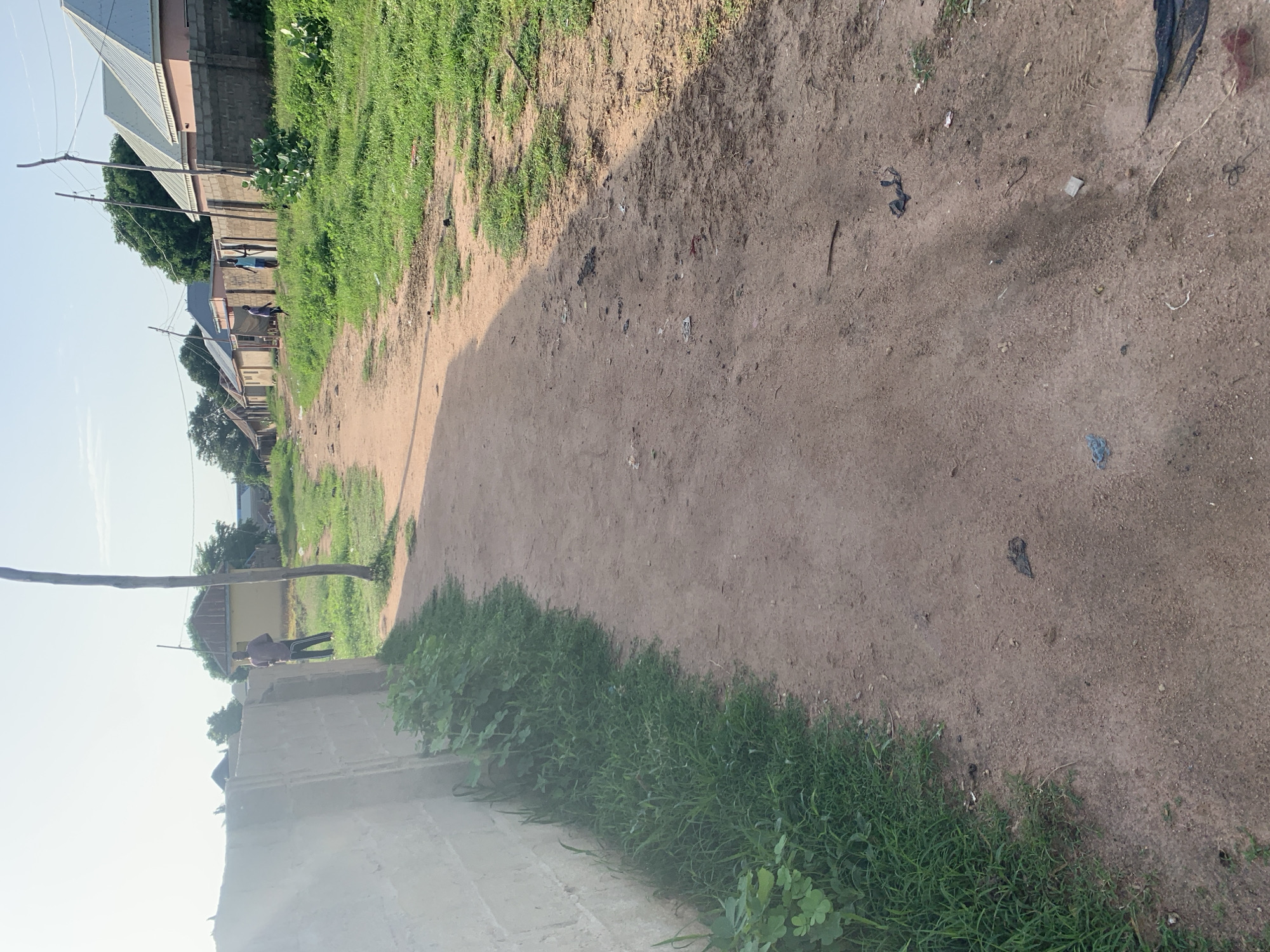 The image depicts a dirt road in Jalingo, Nigeria, flanked by a concrete block wall on the left and sparse vegetation and residential buildings on the right. The road surface is a mix of sand and soil, with some scattered debris, including plastic bags and other refuse. In the background, several single-story buildings with corrugated metal roofs are visible, interspersed with trees. Two individuals, appearing to be young men, are seen standing on the side of the road in the mid-ground. One is wearing a maroon shirt and dark trousers, and the other is in a light blue shirt and dark shorts. The sky is clear with some haze, suggesting it is daytime, possibly late afternoon due to the angle of the shadows. The overall scene conveys a sense of a residential area in a developing urban environment.
