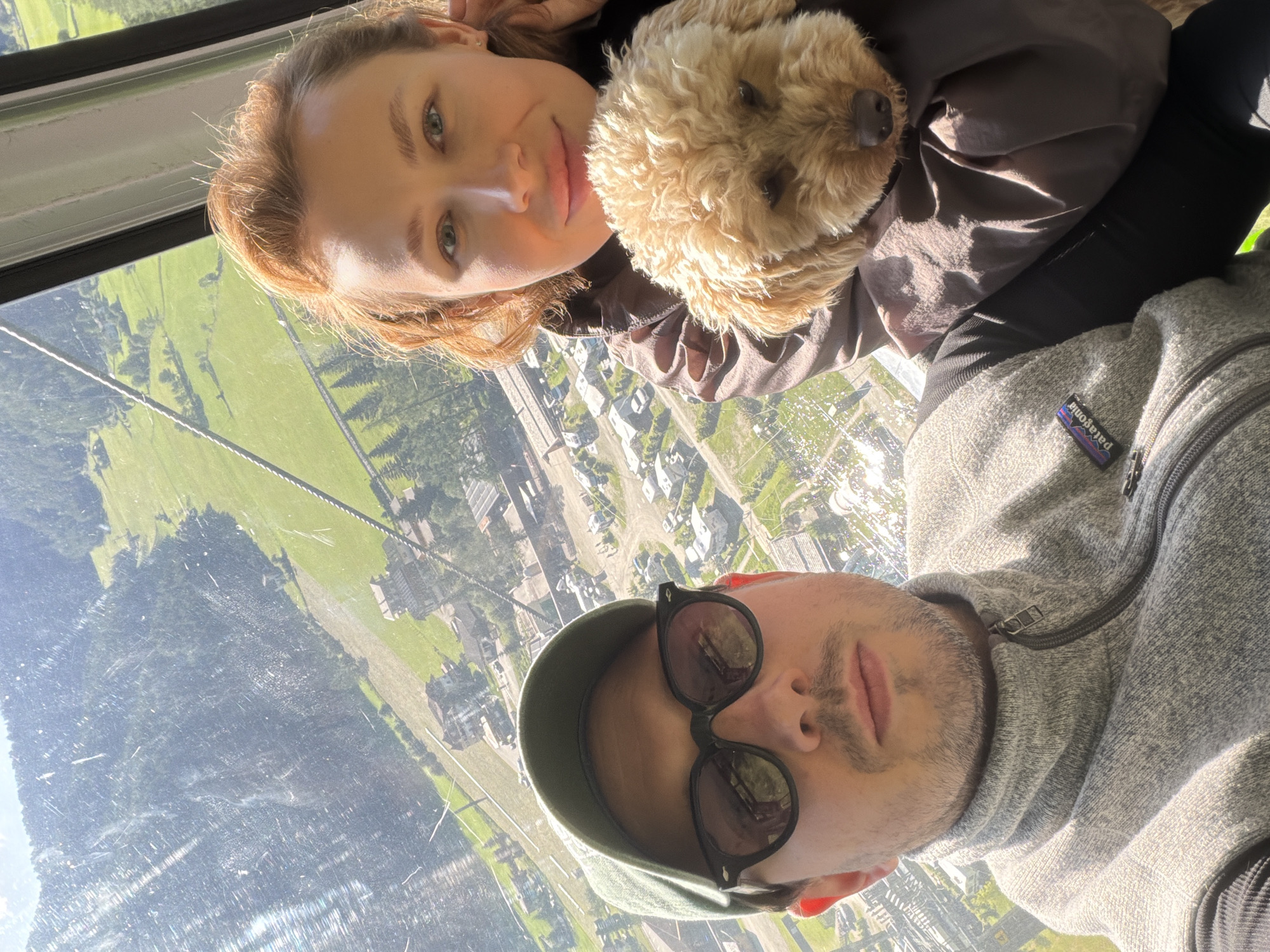 A selfie taken from inside a cable car shows a man and a woman with a dog. The man, on the left, wears a green baseball cap and sunglasses. He has short brown hair and stubble, and is wearing a grey fleece jacket with a Patagonia logo. The woman, on the right, has reddish-brown hair styled up and is looking towards the camera. She is holding a small, light brown poodle-like dog in her arms. They are in a cable car with a window showing a mountainous landscape with green fields, trees, and buildings below. The location is identified as Mühlbach - Rio di Pusteria, Italy. The cable car appears to be moving, as suggested by the slight blur in the background and the reflections on the window. The lighting indicates it is daytime, with sunlight illuminating the scene.