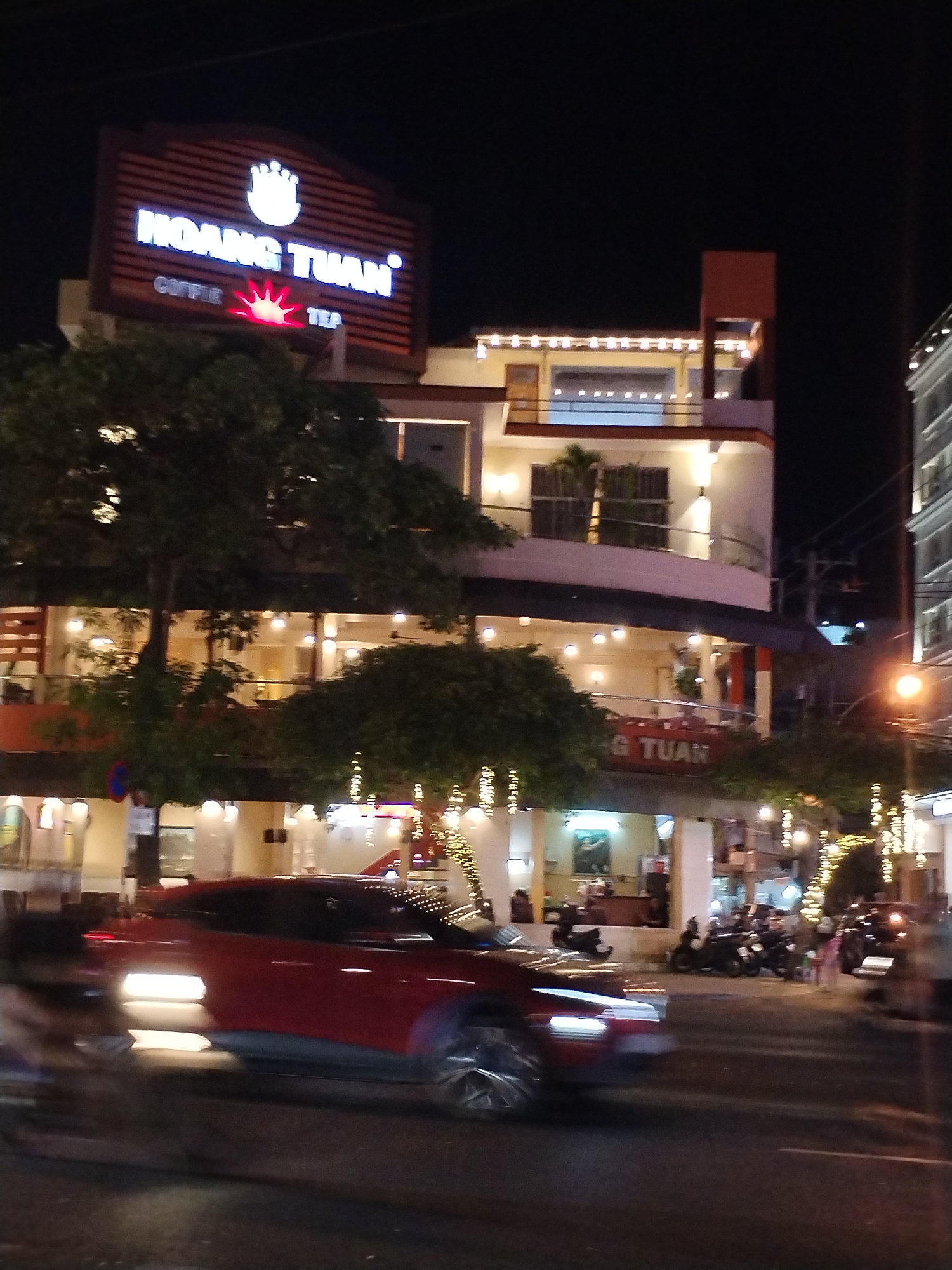 A nighttime image captures a multi-story building in Nha Trang Ward, Vietnam. The building is illuminated by string lights and interior lights, suggesting activity. A large sign reading "HOANG TUAN" is visible atop the building. Trees are present near the base of the building, partially obscuring the ground level. Cars and motorcycles are parked along the street. People are visible on the sidewalk. A red car is moving along the street.