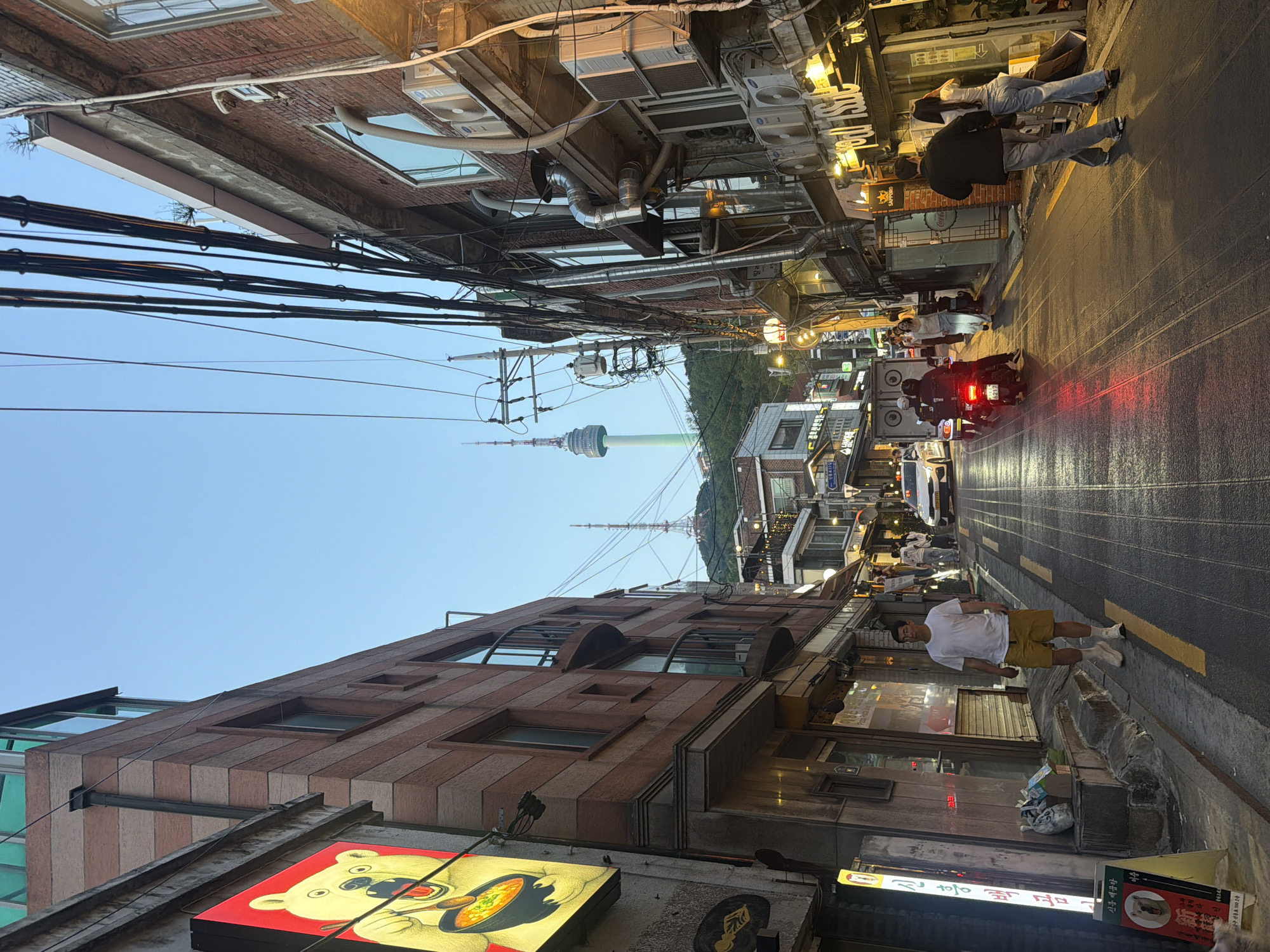 This image depicts a street scene in Seoul, South Korea, recognizable by the Seoul Tower in the background. The view looks along a wet asphalt street flanked by buildings. Electrical wires crisscross overhead. On the right side of the street, two people stand near a storefront, and further down, a person rides a moped with large speakers attached. Several pedestrians are walking along the street. On the left, a man in a white shirt and yellow shorts is walking toward the camera. Visible store signage includes a cartoon bear, implying a restaurant or business of that nature. The building on the left has a facade of horizontally striped bricks. The sky is light blue.
