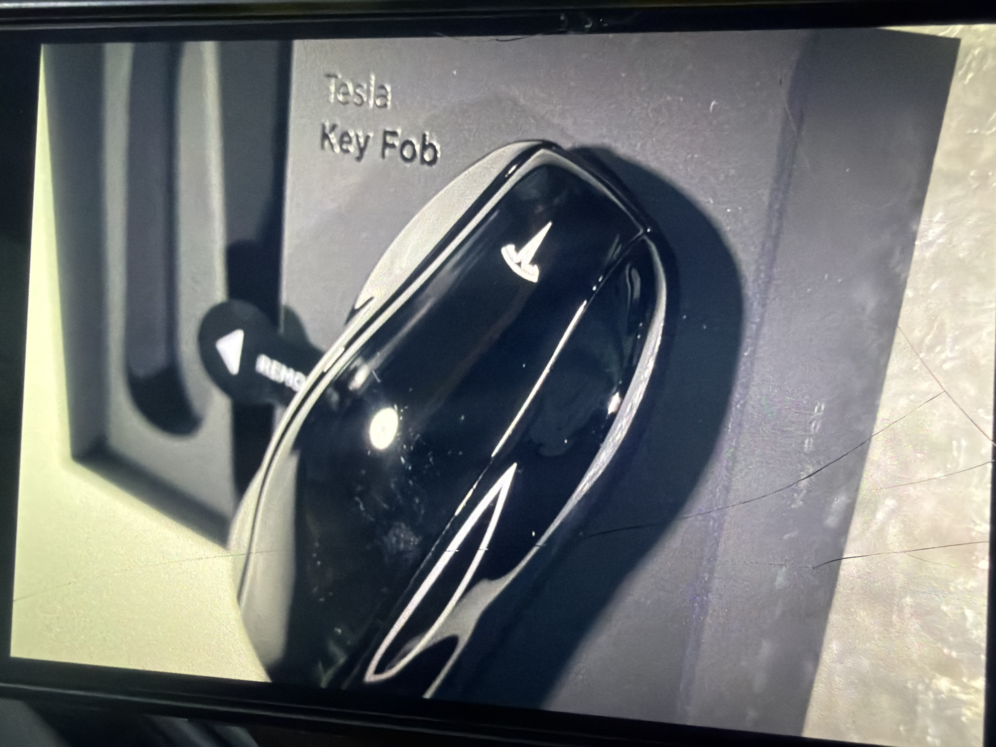 The image shows a black Tesla key fob placed on a dark gray textured surface, which appears to be the packaging for the key fob. The key fob is sleek and has a metallic finish. A small triangular-shaped accessory, likely a key card, is partially visible next to the key fob, with the word "REMOVE" printed on it. The words "Tesla" and "Key Fob" are embossed on the packaging. The scene is brightly lit, and the image is taken from a close-up perspective, with the key fob dominating the foreground. The background is out of focus. The location of Bukuru, Nigeria, is not depicted in the image.