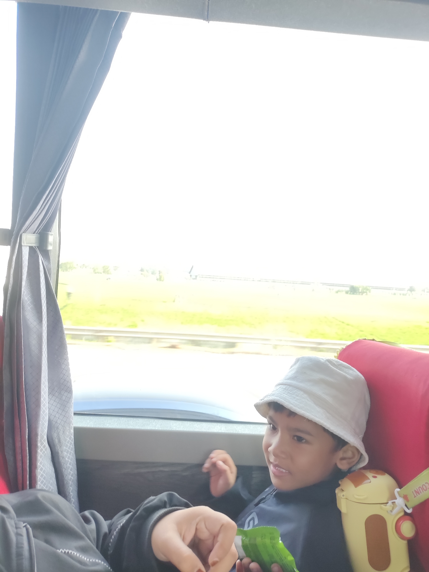 A young boy wearing a white bucket hat and a dark blue shirt sits by a window, looking out at a blurry green landscape. Next to him, a yellow and brown cartoon-themed cup rests on a red cushion. The boy is holding a green object in his right hand. A gray curtain hangs on the left side of the frame. The scene appears to be inside a vehicle, possibly a bus or train, due to the window and interior seating. The bright light suggests it is daytime.