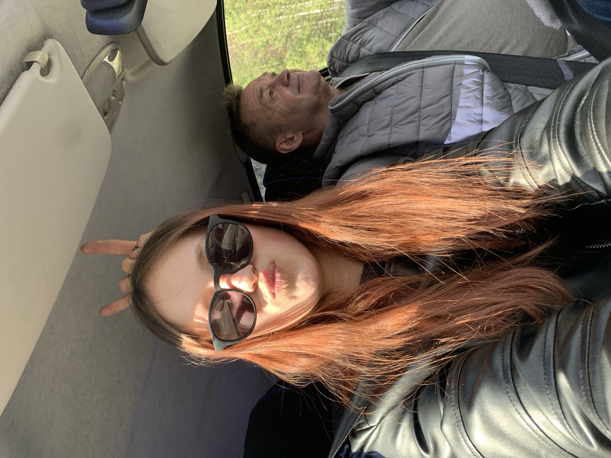 This image, rotated 90 degrees counter-clockwise, captures a playful moment inside a vehicle, likely a car or bus.
In the foreground, a young woman with long, flowing reddish-brown hair poses for a selfie. She wears black sunglasses and a black leather jacket. Her left hand is raised near her head, forming a "rock on" hand gesture with extended index and pinky fingers. Her lips are slightly pursed in a pout, and her face is illuminated by strong sunlight, creating bright highlights and shadows.
Behind her, an older man is seated, wearing a grey quilted jacket. He looks upward and slightly to his left, appearing to gaze out of a window. His expression is calm and contemplative. Through the vehicle's window, green trees and foliage are visible, indicating a natural, possibly forested or rural, outdoor environment, consistent with areas around Chaikovsky, Russia.
The vehicle's interior features a light grey headliner, a sun visor, and an overhead light panel. The strong sunlight suggests a clear, sunny day. There is no visible text or branding within the image. The scene conveys a sense of a casual journey or road trip.