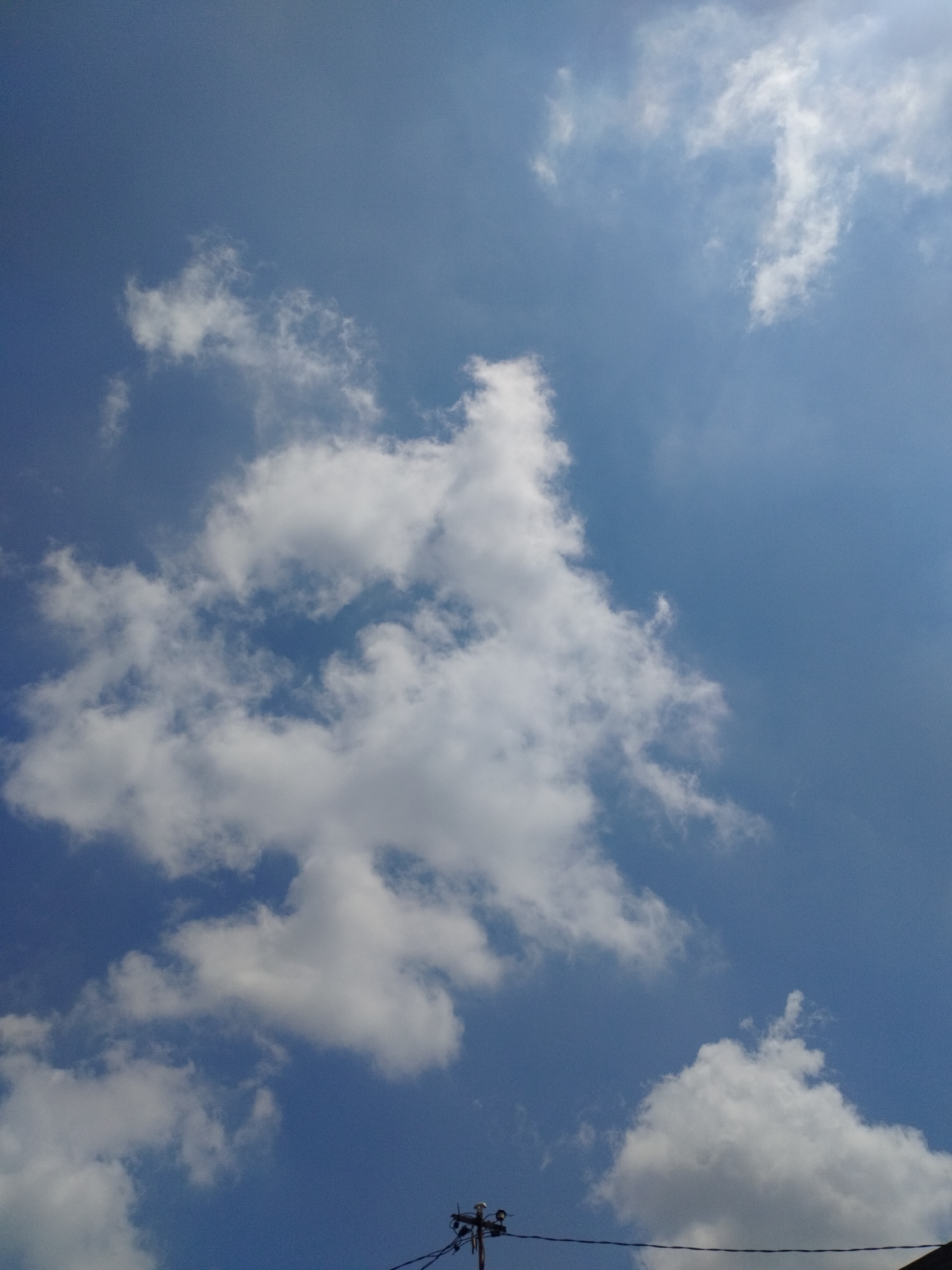 The image shows a bright blue sky with scattered white clouds. A power pole with wires is visible at the bottom of the image. The scene appears to be outdoors, likely in an urban or suburban area, given the presence of the power pole. The lighting suggests it is daytime, with the sun likely overhead and partially obscured by the clouds, casting some shadows. There are no people or animals visible in the frame. The environment is dominated by the sky and clouds, with a man-made structure at the periphery.