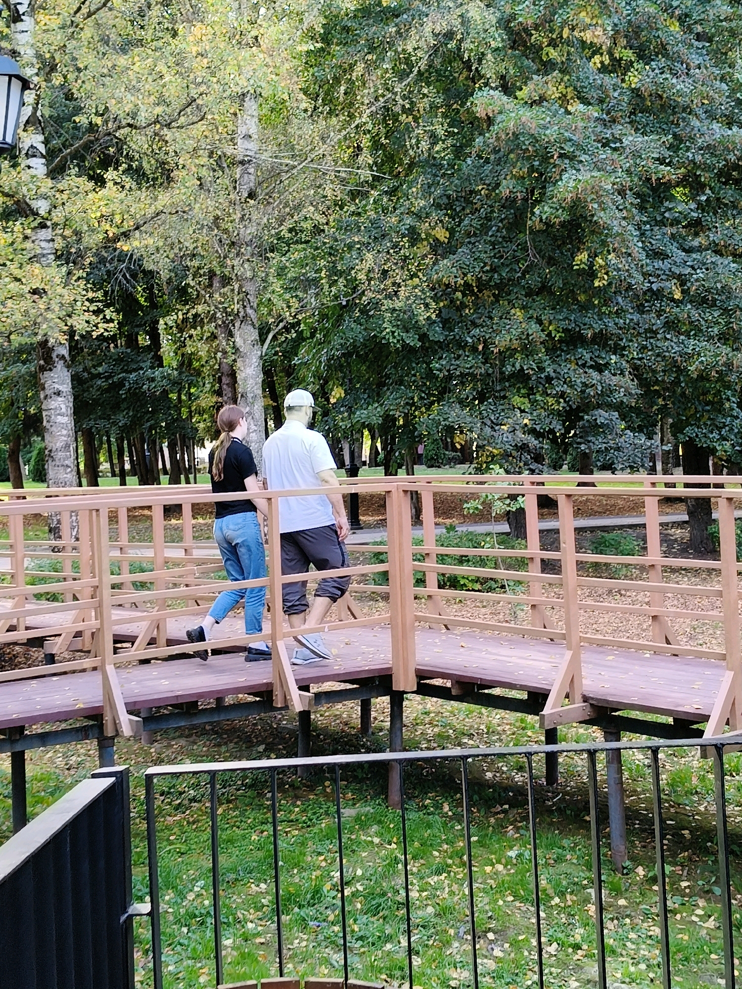 The image depicts two individuals walking across a wooden footbridge in a park-like setting. The male, wearing a white t-shirt, a light-colored baseball cap, and grey shorts, is on the right side of the bridge. The female, wearing a black t-shirt and blue jeans, is on the left. They are walking away from the viewer towards the right side of the frame. The footbridge, constructed from wood with light-colored railings, spans over a grassy area with scattered fallen leaves, suggesting an autumn season. Trees with lush green and some yellowing foliage form a dense backdrop, with birch trees and other deciduous varieties visible. A black metal fence is in the foreground, partially obscuring the view of the ground beneath the footbridge. The lighting suggests it is daytime, possibly late afternoon, with soft shadows cast by the trees and the bridge structure. The scene appears to be in a public park, likely in Naro-Fominsk, Russia, given the typical urban park aesthetic.