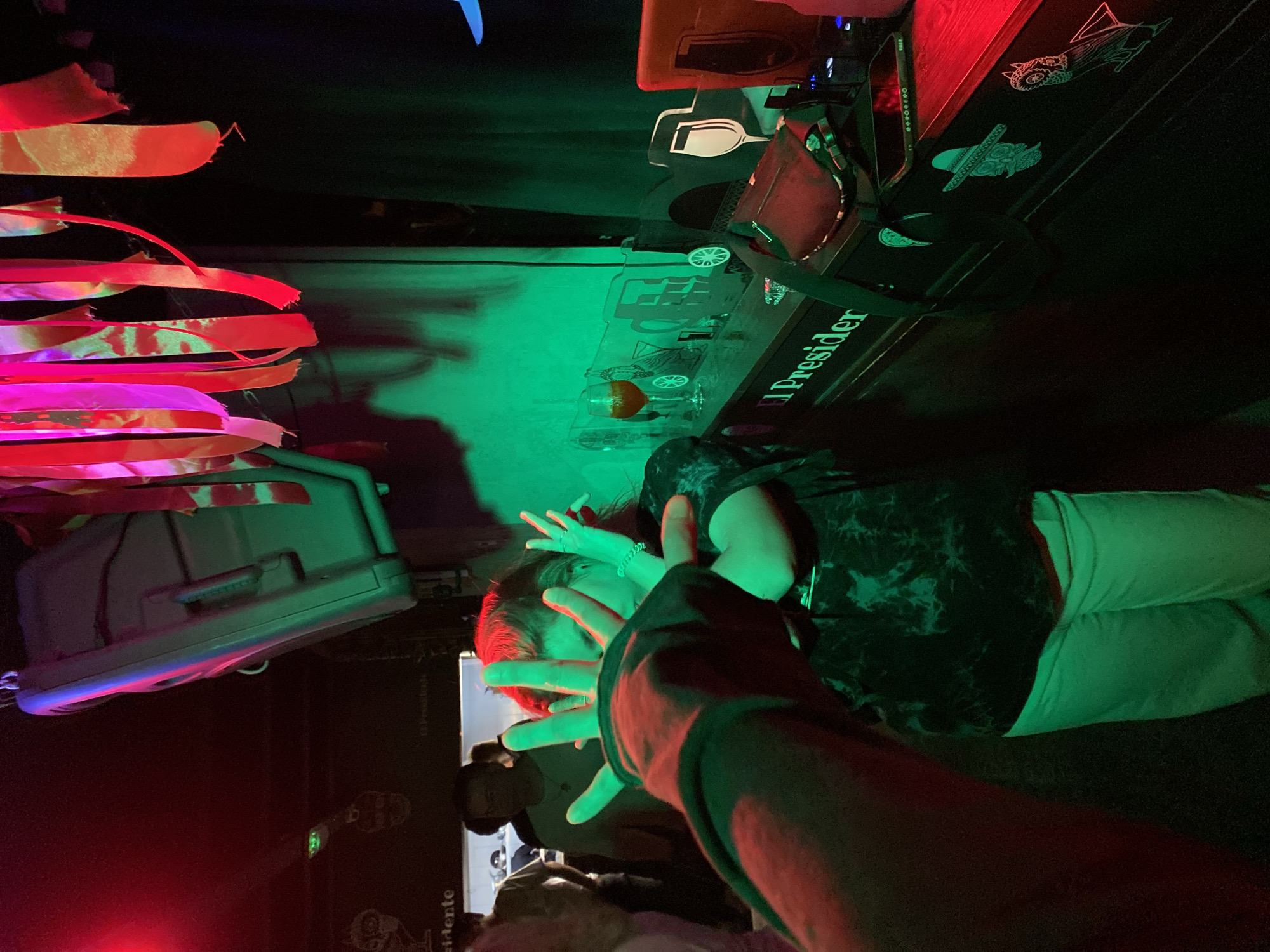 A nightclub scene is captured under red and green lighting, showing a woman covering her face with her hands, which are in the foreground. She is wearing a dark patterned shirt and light-colored pants. A second person is partially visible behind her. The "President" sign is visible on a shelf or counter. Decoration includes streamers and a piece of luggage. The location cannot be determined from the provided image.