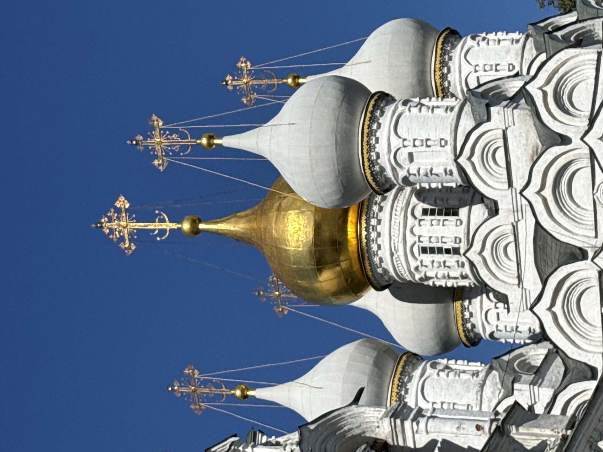 The image shows a close-up view of the domes and crosses of the Archangel Cathedral, located in the Moscow Kremlin, Russia. The cathedral's white, ornate architecture is contrasted against a clear blue sky. The domes are topped with golden crosses, with one prominent golden dome in the center. The detailing on the structure, including decorative moldings and small windows, is visible. The image is a still shot, with no people or animals present. The lighting suggests it is daytime, likely with bright sunlight.