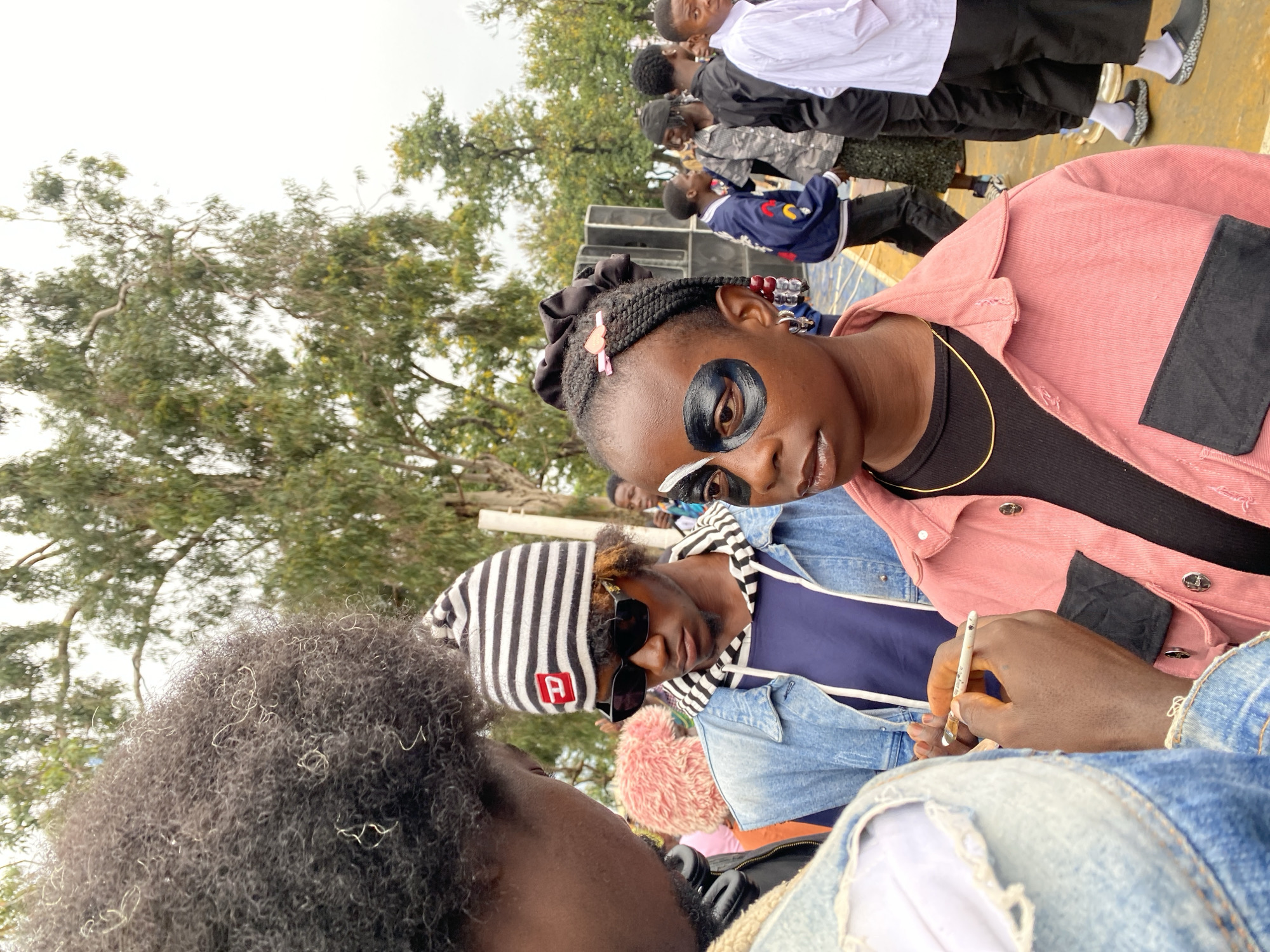 A young Black person with braided hair and decorative beads is having their face painted, with black and white paint applied around one eye, at an outdoor event in Bar Jirgi Summa, Nigeria. They are wearing a pink jacket over a black top and a gold chain. To their left, another Black person, wearing a denim jacket and a striped beanie, is observing the face painting. In the background, other people are visible, as well as trees and what appears to be a sound system. The ground is a yellowish-orange surface, possibly painted concrete or a similar material, indicating an event space or recreational area. The sky is overcast.