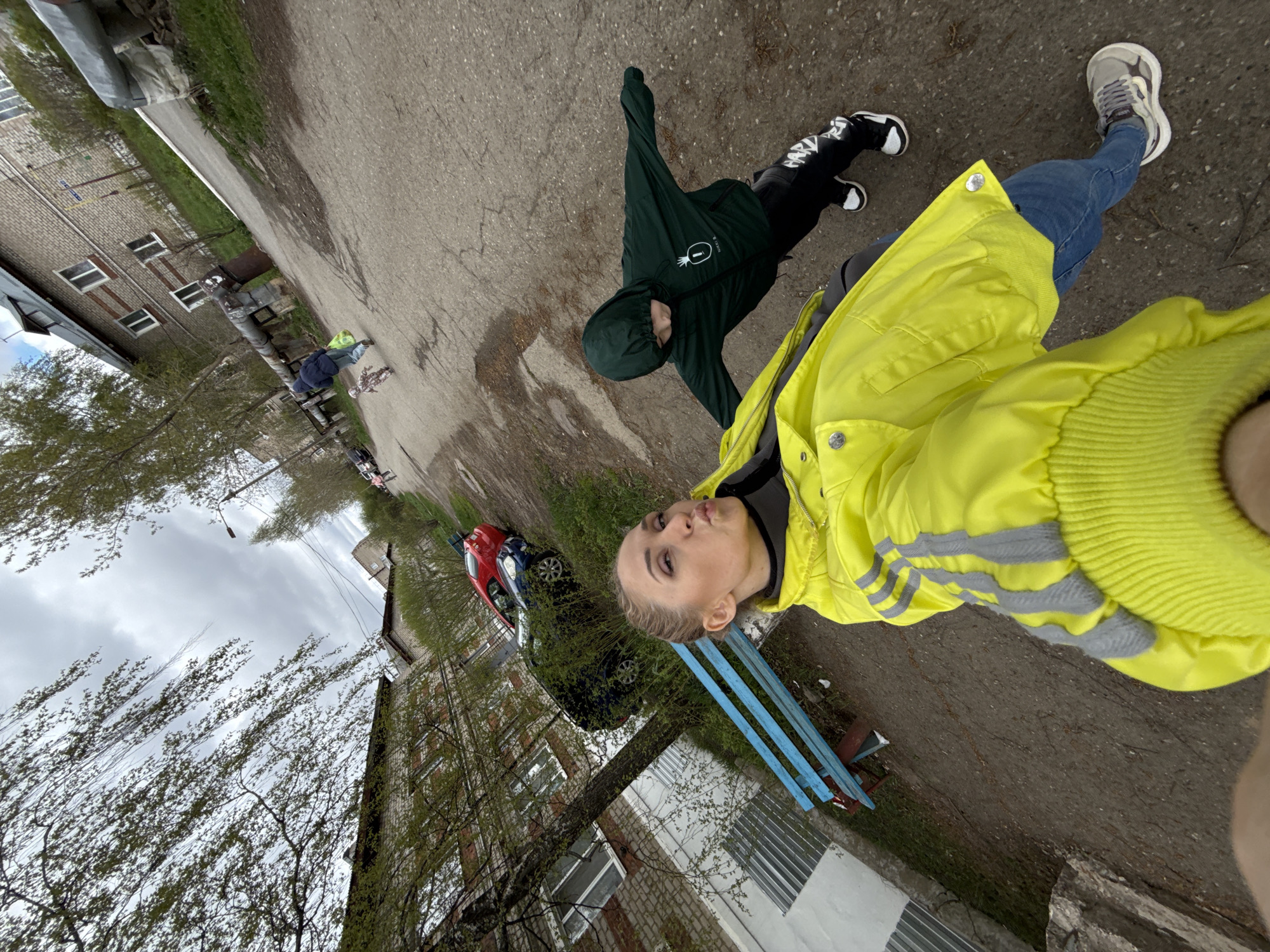 A medium shot captures a person wearing a yellow jacket, blue jeans, and grey sneakers, appearing to take a selfie on a street. Next to them is a child in a dark green hooded jacket and black and white pants. The setting includes an asphalt street, grass edges, a blue bench, and parked red and blue cars. In the background, brick apartment buildings are visible under an overcast sky with bare trees. The location is Vereshchagino, Russia.