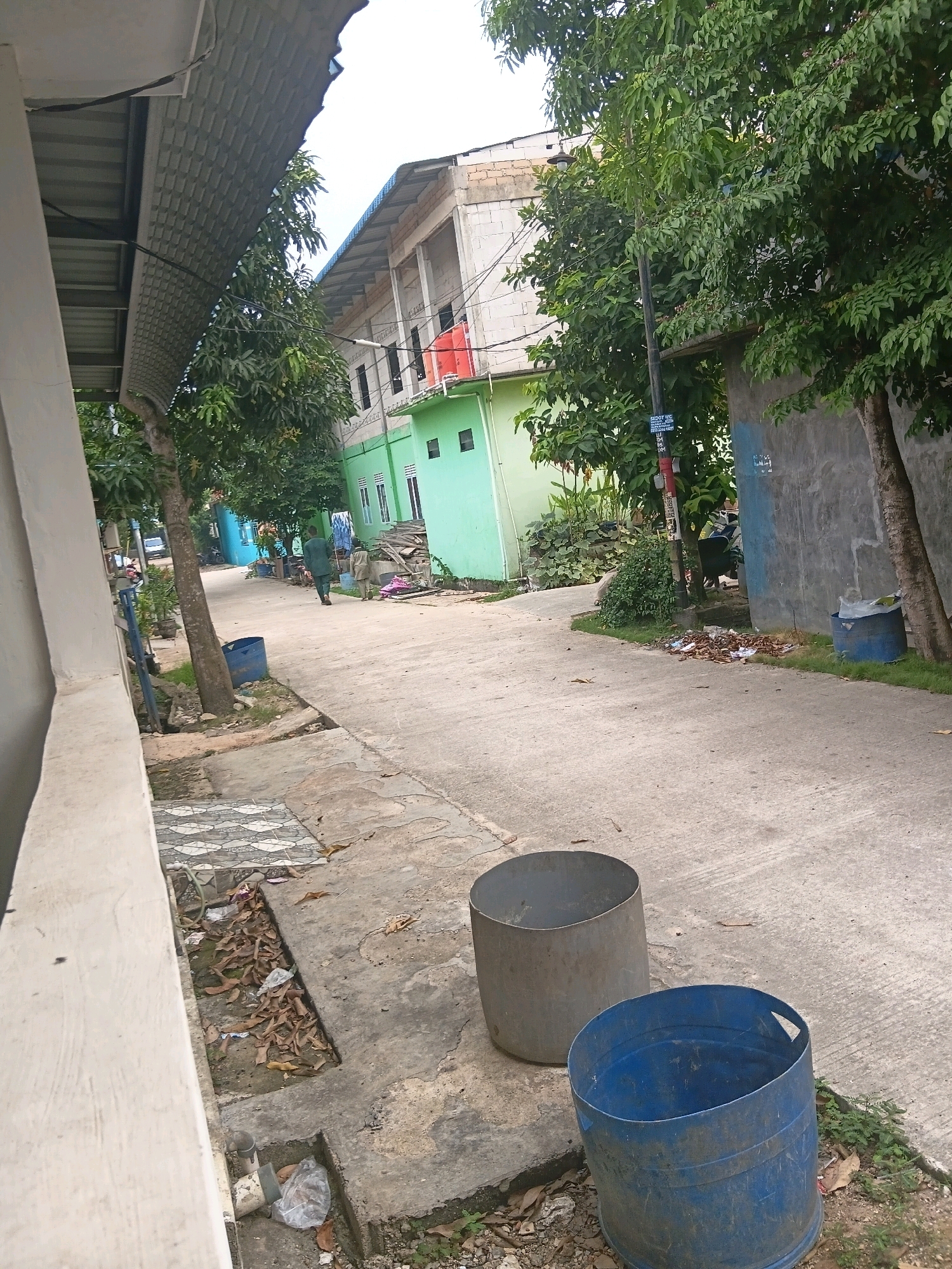 The image depicts a residential street in an urban area, likely in Southeast Asia, with buildings on either side of a paved road. A two-story building painted green and white with red accents is visible in the background. Trees with dense green foliage line the street, providing shade. In the foreground, two large cylindrical bins, one gray and one blue, are placed on the pavement. A person in a green shirt is walking away from the camera down the street, towards the buildings. Scattered leaves and debris are present on the ground, suggesting an outdoor environment. The sky is overcast, indicating a cloudy day. The overall scene portrays a typical street view in a populated area, with elements of everyday life such as buildings, trees, and waste receptacles.