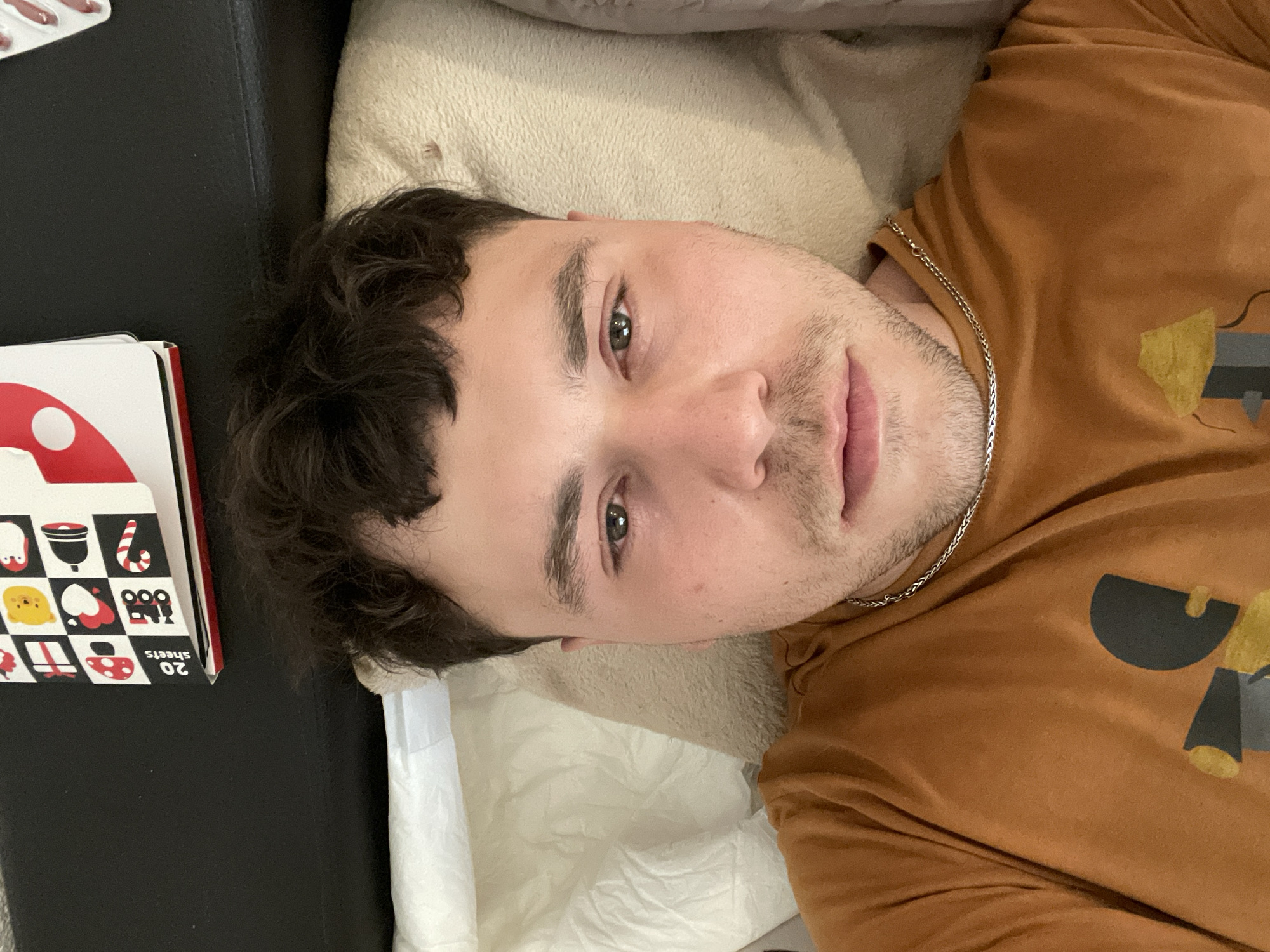 A person lies on a bed in an indoor setting. The individual, likely male, has short brown hair, light skin, and is wearing a brown t-shirt with a design and a silver chain. The person is positioned on a white pillow or cushion, which rests on a lighter-colored upholstered surface. Beside the person is a box labeled "20 sheets" with graphics of various objects. The location in the city of Bar, Montenegro cannot be determined from the image.