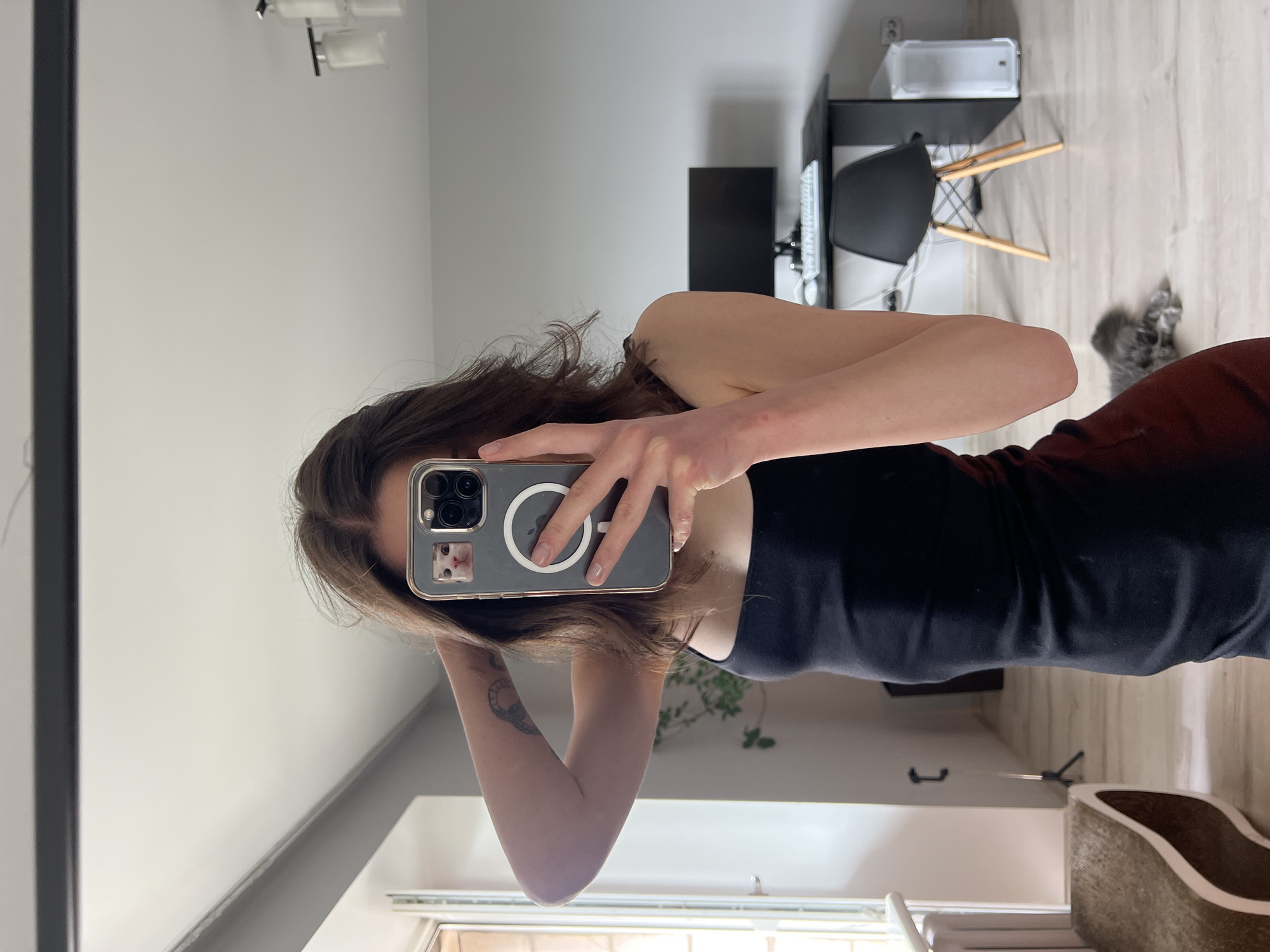 A young woman with medium-length brown hair takes a mirror selfie in what appears to be a modern apartment living room or home office. She is wearing a black tank top. Her left arm is raised, with her hand touching the back of her head. She is holding a smartphone with a case featuring a picture of a cat. In the background, a desk with a computer monitor and keyboard is visible, along with a black chair and a white computer tower. A window is to her left, and a cat lies on the floor in the lower right portion of the image. The room is well-lit, suggesting it is daytime. The presence of a computer setup and the general decor indicate a contemporary living space. The image does not provide specific clues that would place it in Ternopil, Ukraine, or any other particular city.