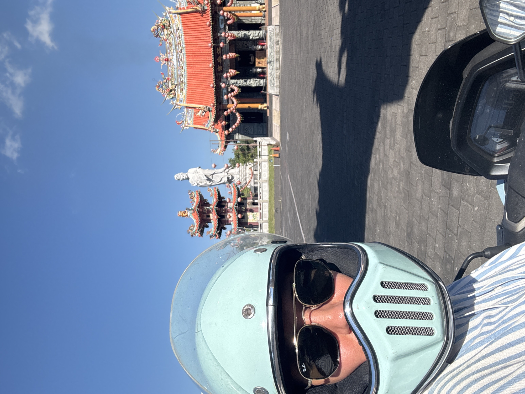 The image captures a person wearing a light blue helmet and sunglasses, seemingly on a motor scooter. In the background, there is a traditional Asian building with red roofs and ornate decorations, along with a large white statue. The setting appears to be a paved area in Denpasar, Indonesia, based on the architectural style of the building and the presence of the statue, which could potentially be a religious monument. The sky is clear and blue, suggesting a sunny day. The angle of the shot suggests it was taken from the first-person perspective of the person wearing the helmet.