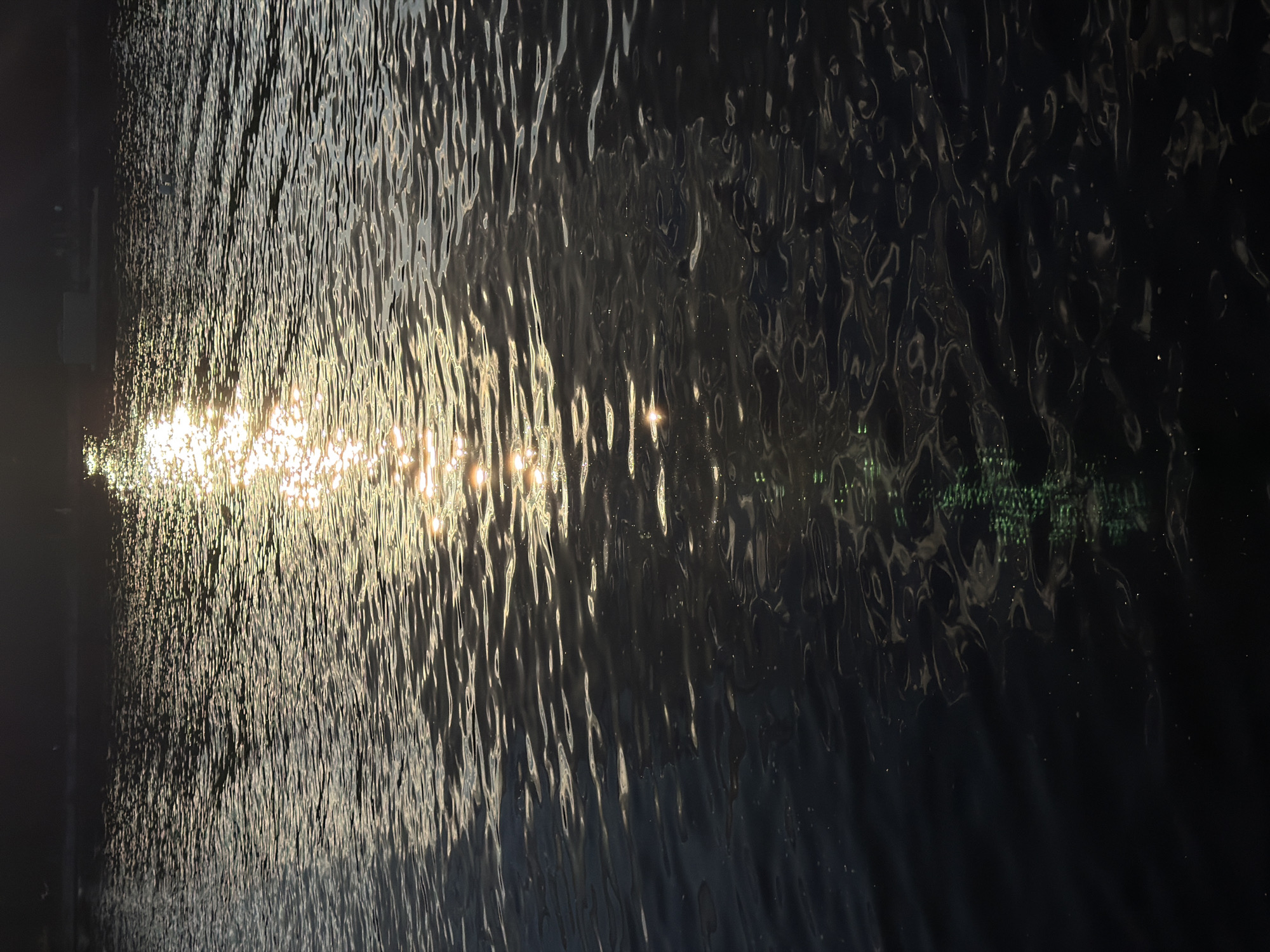 The image depicts a body of water, likely a river or lake, with sunlight reflecting off its surface. The water appears dark, with ripples creating patterns of light and shadow. In the upper portion of the image, the sun's reflection is bright and elongated, suggesting it is low in the sky, possibly during sunrise or sunset. A dark, indistinct shoreline is visible at the top edge. The overall scene is natural and tranquil.
This scene could be located on the Iset River in Yekaterinburg, Russia, particularly in an area where the water is calm and the sun's reflection is visible.