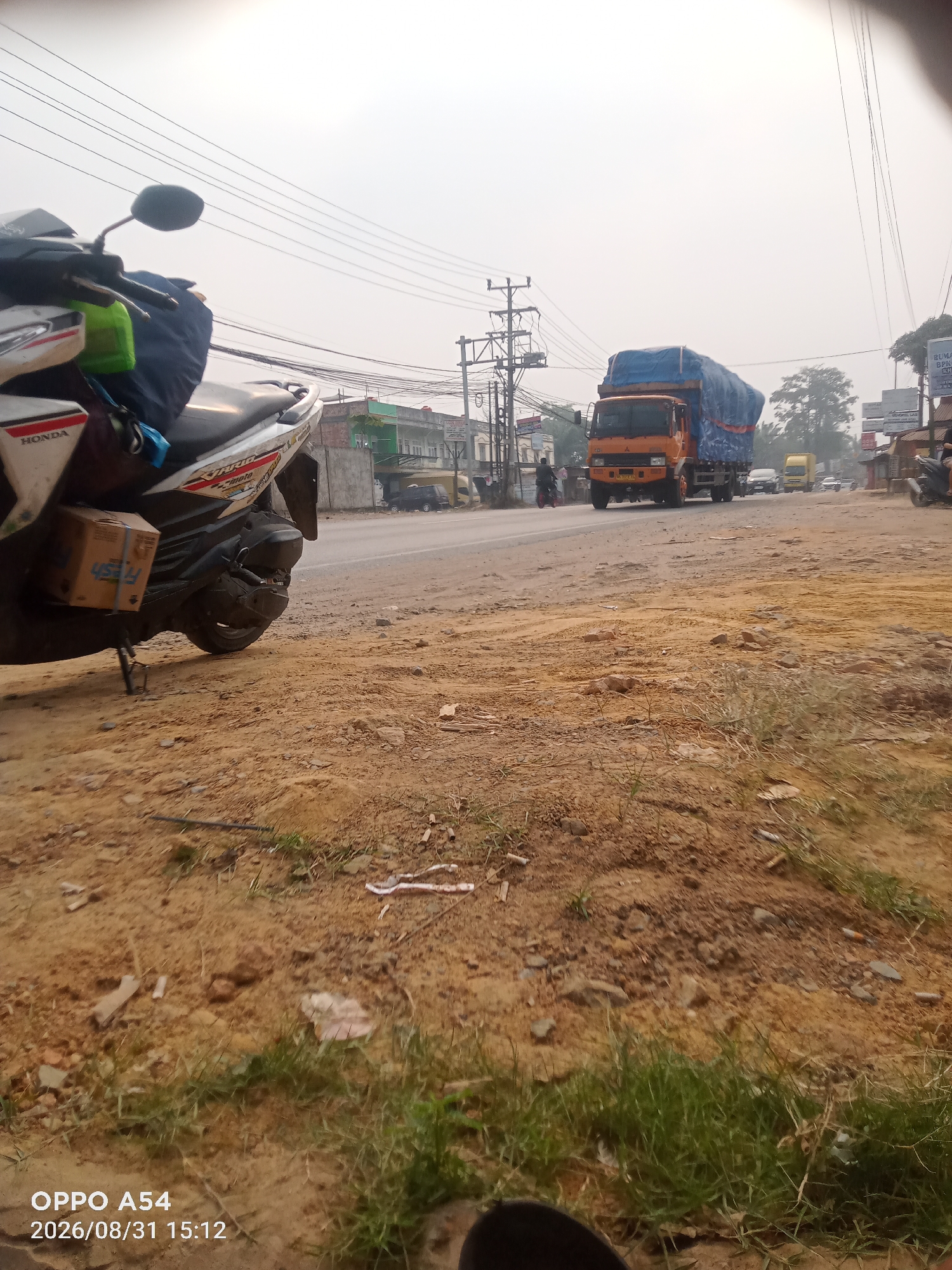 The image depicts a roadside scene in Jambi, Indonesia, captured on August 31, 2026, at 15:12. A white and red Honda motorcycle is parked on the right side of the frame, with a brown cardboard box secured to its side. The motorcycle is parked on a dirt and gravel shoulder, with sparse green grass growing in patches. In the background, a road runs horizontally across the image. Several vehicles are visible on the road, including an orange truck with a blue tarp covering its cargo, a yellow van, and other cars. Buildings with green and white facades line the right side of the road. Overhead, power lines crisscross the sky. The overall atmosphere appears hazy or overcast. The timestamp and device information "OPPO A54 2026/08/31 15:12" are present in the bottom left corner.