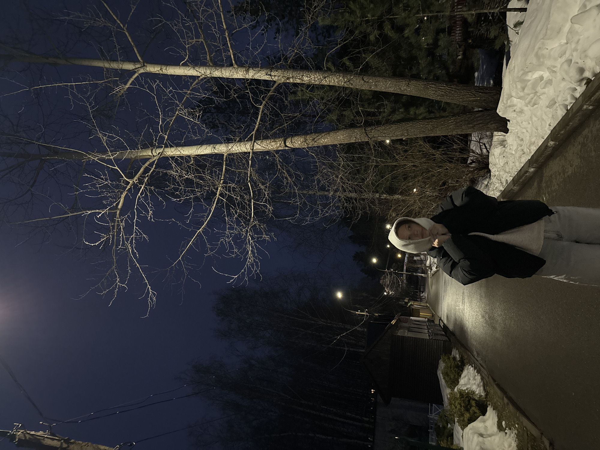A woman stands on a paved pathway at night in Chaikovsky, Russia. Bare trees are visible in the background against a dark blue sky. Streetlights illuminate the area. Snow is piled on the side of the path. Houses are visible in the distance. The woman is wearing a dark coat and a hooded jacket.