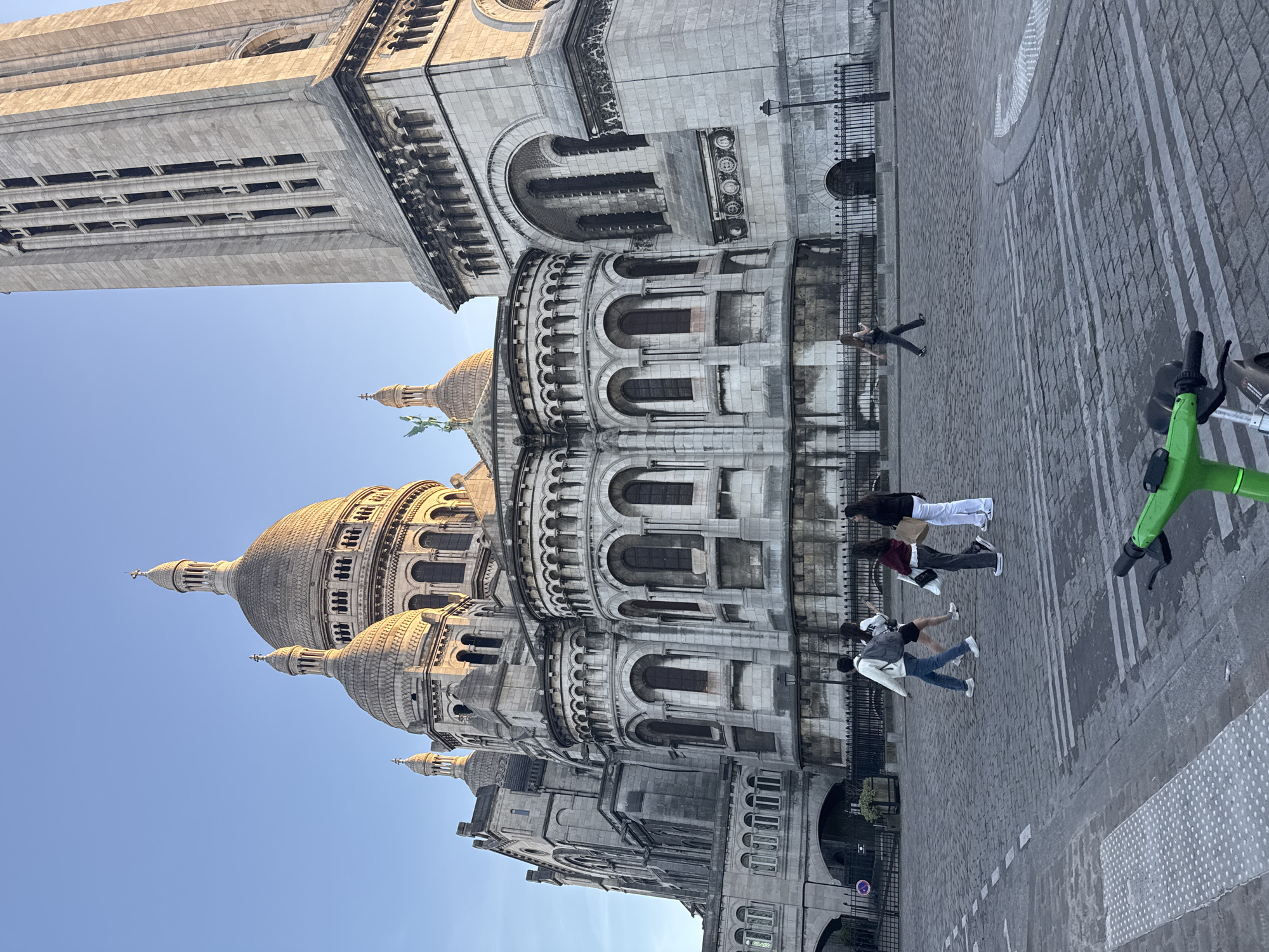 The image depicts the Sacré-Cœur Basilica in Paris, France, viewed from a low angle looking upwards. The sky is clear and blue. The basilica's architecture is prominent, showcasing its domes, arches, and details. In the foreground, on the paved street, there are several people standing and walking, and a green electric scooter is parked. One person is lying on the pavement, perhaps posing for a photo. The scene appears to be taken during the daytime.