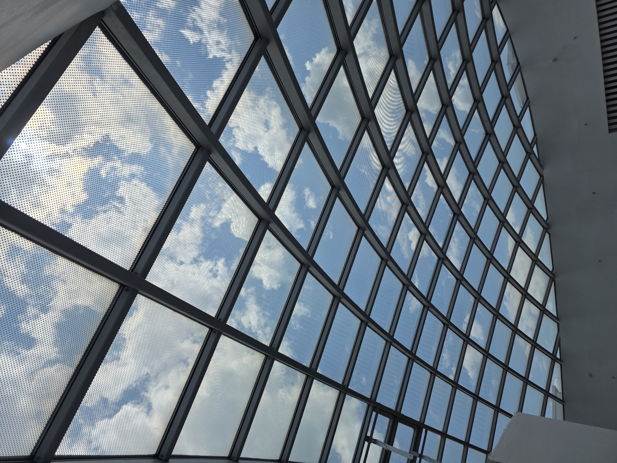 The image shows a section of the glass roof of the Mall Plovdiv in Plovdiv, Bulgaria. The roof is constructed of multiple small, rectangular glass panels held together by a grid of grey metal supports. Each panel appears to have a dotted pattern applied to its surface. Through the glass, a view of a bright blue sky with scattered white clouds is visible. The angle of the shot is looking upwards and towards the side of the roof, creating a perspective effect where the panels appear to get smaller in the distance. Part of the interior architecture, including white walls and ceiling, are visible in the upper right corner of the image.