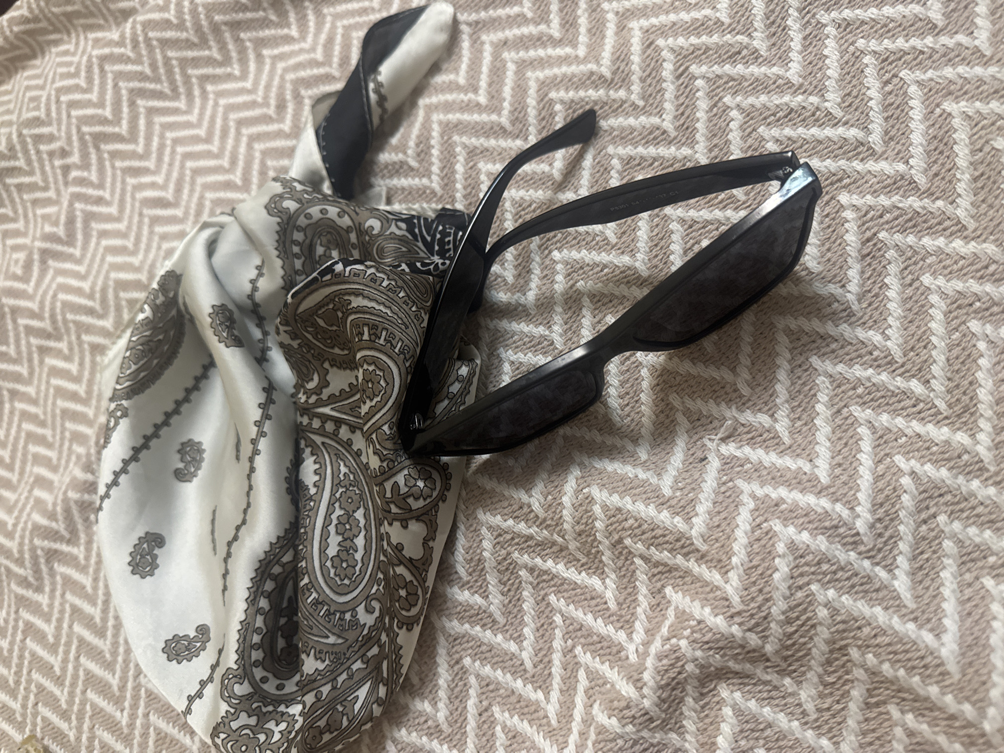 The image shows a close-up, overhead view of a black pair of sunglasses and a patterned silk scarf with paisley designs, resting on a textured surface. The surface appears to be a blanket or throw with a zig-zag pattern in light brown and white. The sunglasses are partially open, with one arm extended and the other resting on the scarf. The scarf is gathered and draped, with its intricate pattern of paisleys visible in shades of black, grey, and white against the cream-colored fabric. The lighting is soft and natural, creating gentle shadows. There are no people visible in the image, and no specific location in Omsk, Russia can be identified from the provided visual information.