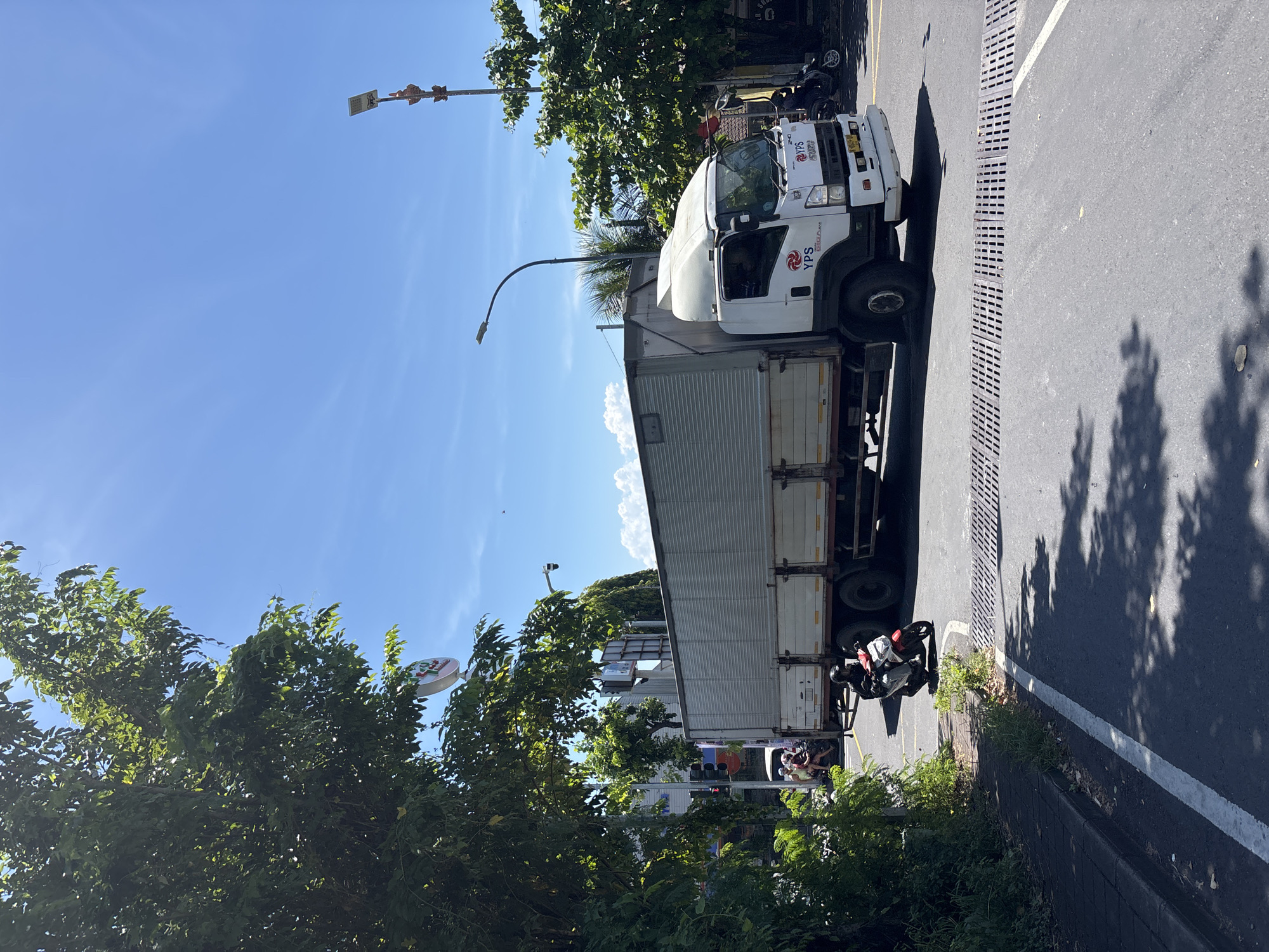 The image shows a truck parked on the side of a road in Nusa Dua, Indonesia. The truck is white with a container trailer. The brand "YPS" is visible on the side of the cab. A motorcycle is parked behind the truck. Trees and shrubs line the side of the road. A street light and signs are also visible. The sky is blue with some clouds. The road is asphalt with a white line marking the edge.