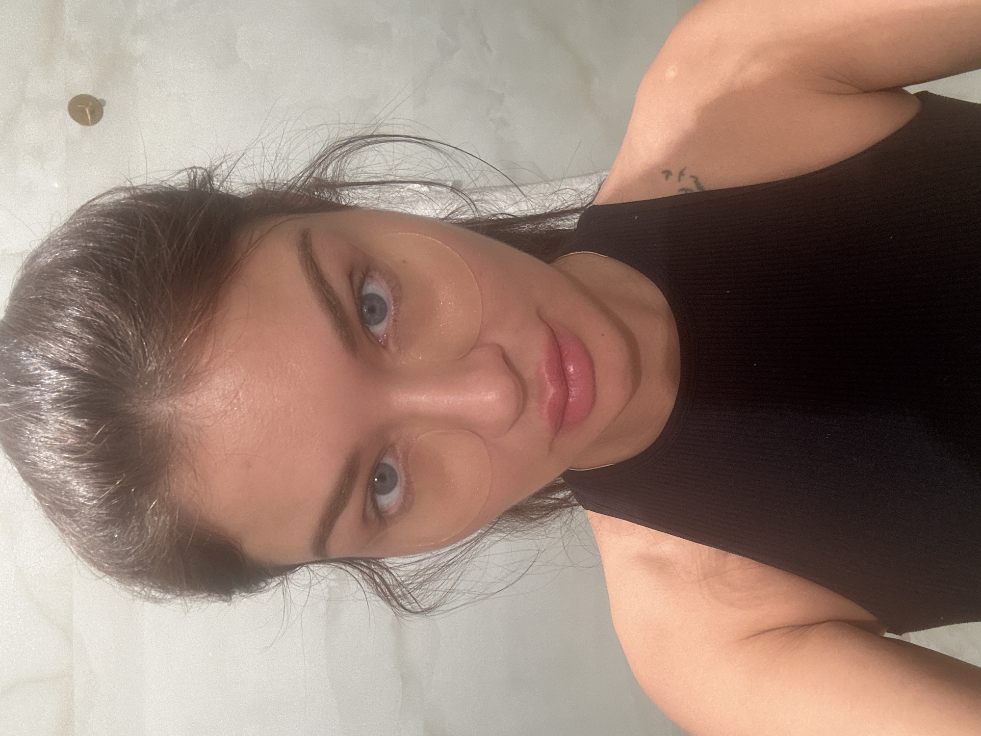 A selfie shot shows a woman with dark hair and blue eyes. She has under-eye patches applied to her face. She is wearing a black sleeveless top. Part of a tattoo is visible on her shoulder. The background appears to be a white marble or tiled wall. The image does not provide sufficient detail to accurately place it in a specific location in Moscow, Russia.