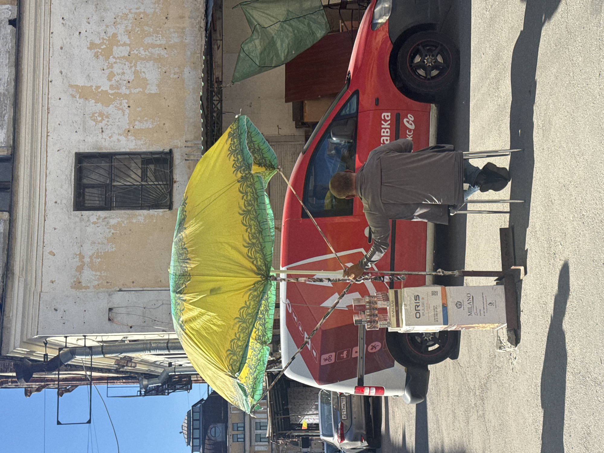 The image depicts a street scene in Saint Petersburg, Russia, on a sunny day. A man is seated on a small, metal stool in front of a red van, holding the pole of a large, open yellow umbrella adorned with a green palm-tree motif. The umbrella provides shade over the man and a display of stacked boxes, likely of tobacco products. The boxes have visible branding, including "BARCLAY," "ORIS PULSE," and "MILANO COFFEE." The red van has white lettering on its side, including "ПЛАТЕВКА" and "КС Go". In the background, there's an old building with textured, peeling plaster and a barred window. To the left, a grey car is parked, and the roof of another building with a distinctive dome is visible. The ground is asphalt, with distinct shadows cast by the objects and people.