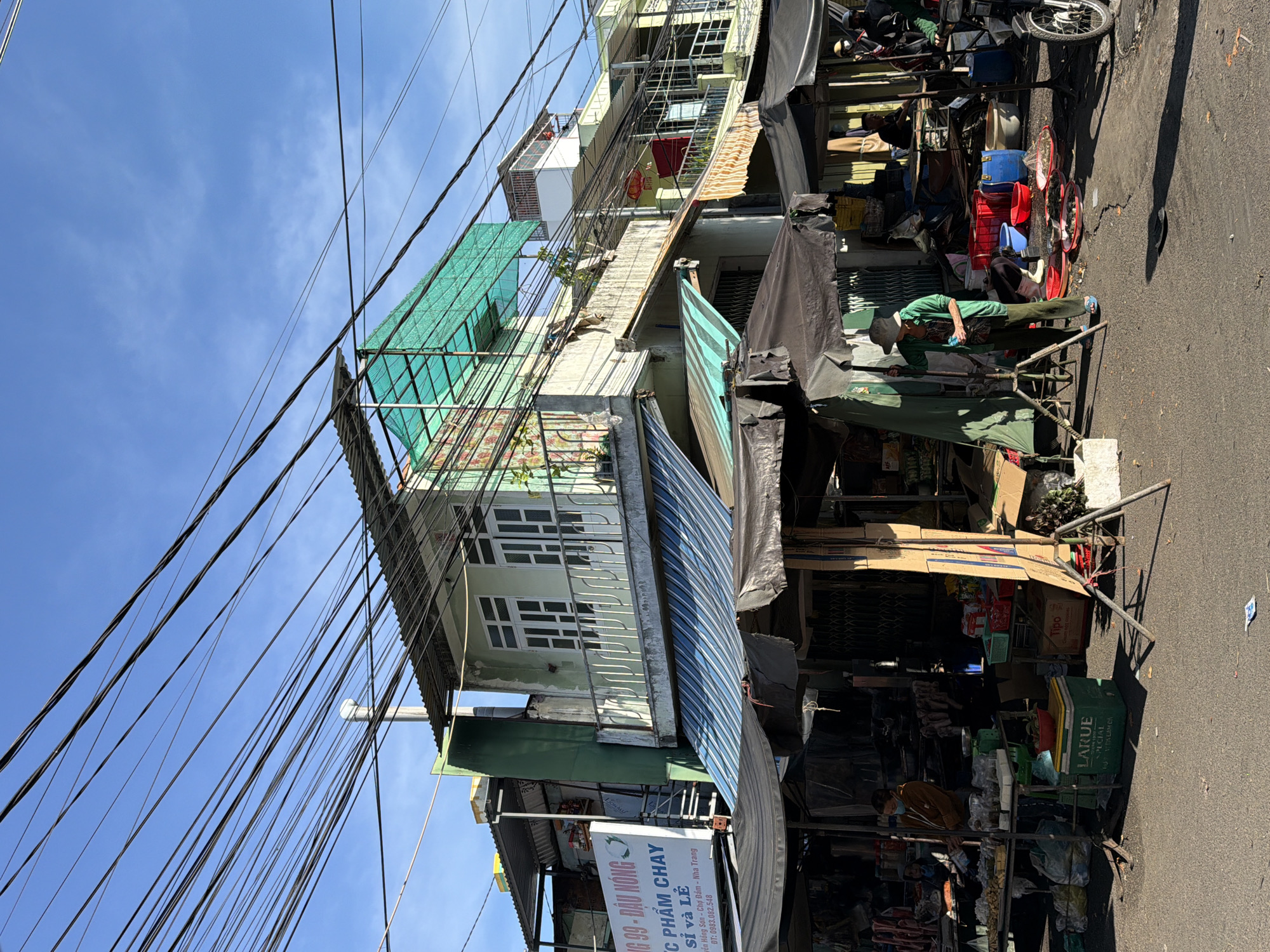 The image depicts a street market scene in Nha Trang, Vietnam, characterized by a dense network of overhead electrical wires against a clear blue sky. In the foreground, several market stalls are visible, covered by dark tarps and awnings, some with blue and white striped fabric. Various goods are displayed on tables, including produce, packaged items, and containers. One stall features a green cooler with a "LARUE" label and a red bowl on top.
In the middle ground, a person wearing a green long-sleeved shirt, dark pants, and a hat is standing next to a tripod, appearing to be setting up equipment or observing. Other individuals are seen in the background, some appearing to be vendors or customers. Behind the market stalls, buildings with multiple stories are visible, some with balconies and windows. The architecture suggests a mix of residential and commercial spaces.
The overall environment is that of a bustling outdoor market, likely in a densely populated urban area. The presence of numerous electrical wires is a common sight in Vietnamese cities, indicative of the country's infrastructure. The ground is paved, and scattered debris suggests the ongoing activity of the market. The lighting indicates it is daytime, with shadows cast by the structures and objects.