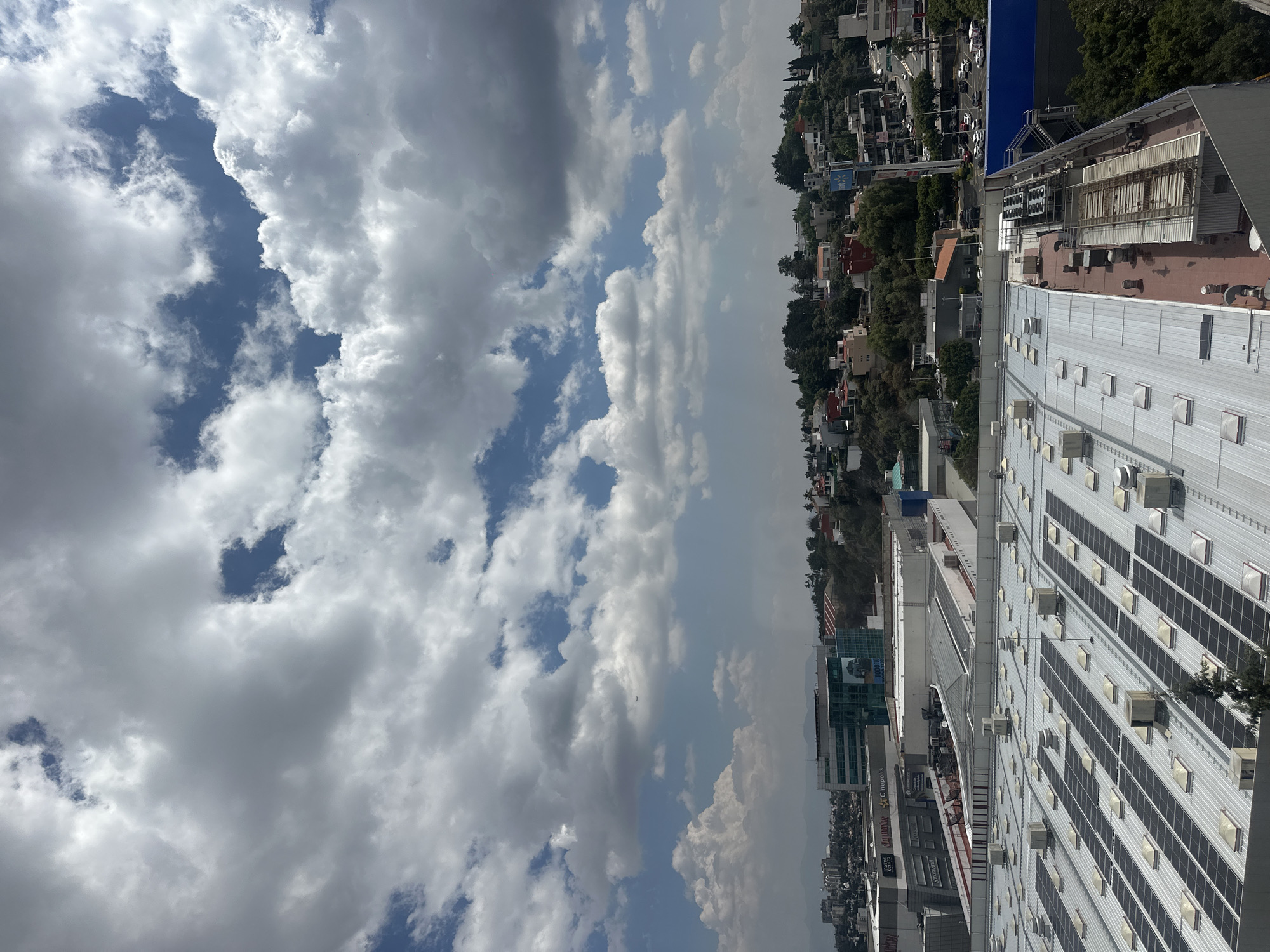 The image shows an elevated view of the city of Huixquilucan de Degollado, Mexico, under a cloudy sky. The perspective is from atop a tall building. A large roof covered with solar panels dominates the lower right portion of the view. The surrounding buildings and cityscape stretch towards the horizon, with a mix of residential and commercial structures visible. Trees dot the landscape, particularly on a hillside in the distance.