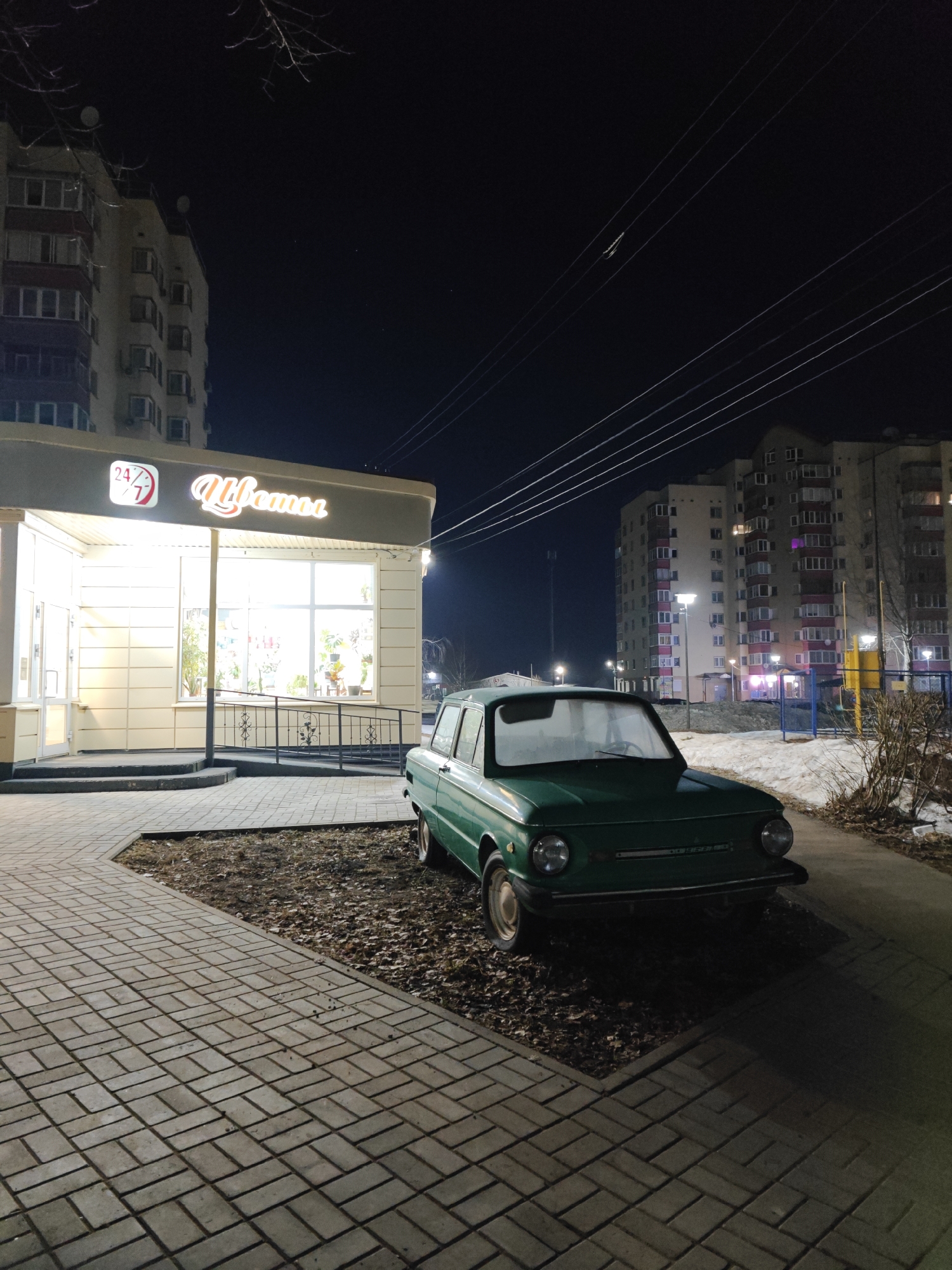 The image depicts a ZAZ-968 car, dark green in color, parked on what appears to be a parking area next to a sidewalk, with bare-leaf mulch around the vehicle. A building with a lit-up sign in Russian, "Цветы" (Flowers), with a 24/7 symbol, indicating a flower shop, is to the left of the car. The building is beige. In the background, there are residential apartment buildings under a night sky. The location is likely Chaikovsky, Russia, given the architectural style and car type. The scene is illuminated by streetlights and the flower shop's lights. Patches of snow are visible behind the car near a playground.