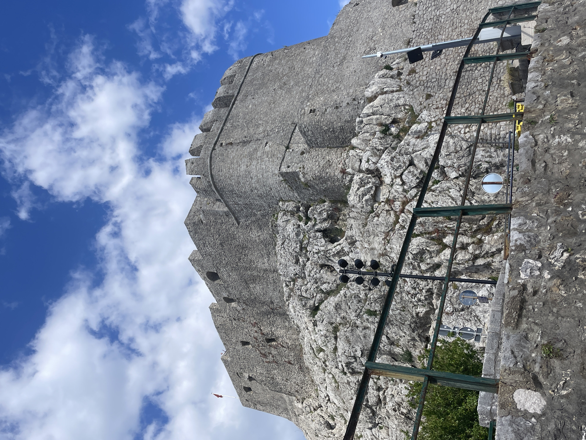 This striking image captures a formidable stone fortress set against a bright blue sky with scattered white clouds, likely in Herceg Novi, Montenegro. The massive grey stone walls of the fortress dominate the left and upper parts of the frame, showcasing ancient construction with irregular, fitted stones. Along the top of the wall are crenellated battlements, hinting at its defensive purpose. A dark, possibly metal, pipe or conduit runs vertically down a section of the wall. On one of the higher sections, a small red flag is visible, fluttering in the breeze.
The fortress is built into and upon a rugged, rocky hillside, with exposed natural rock formations forming its base and extending into the midground on the right. In the foreground and midground, a dark green or black metal framework structure is prominent, angled diagonally across the right side of the image. This structure appears to be a walkway, support system, or part of an old transportation mechanism, with multiple beams. Attached to or near this framework are black multi-lamp spotlights or streetlights.
Further down and behind the metal structure, a circular white sign with a horizontal black bar is clearly visible, possibly indicating "no entry" or a similar restriction. Another similar sign is partially obscured below it. Some green foliage and small trees are visible nestled among the rocks and below the structures.
The scene depicts a sunny daytime, evidenced by the clear blue sky and bright lighting on the fortress and rocks. No people or specific activities are observable, lending a quiet, majestic quality to the historical structure. No discernible text is visible on any signs or objects. The image conveys the imposing scale and historical presence of the fortress within its natural, rugged environment.