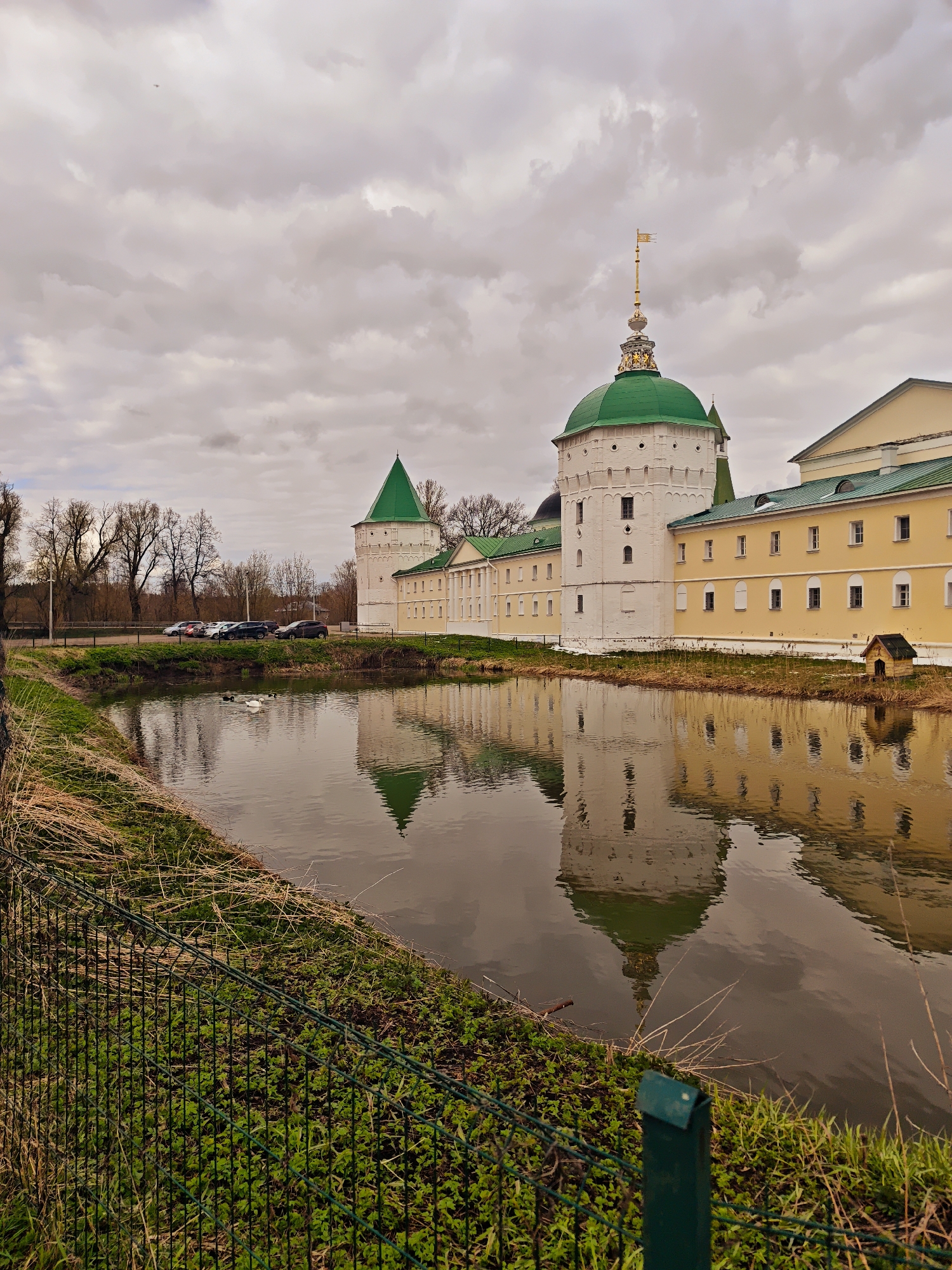 The image displays a picturesque scene featuring a large architectural complex, likely a monastery or fortress, situated beside a tranquil pond in Луговой, Russia. The complex comprises white-walled buildings with prominent green roofs, including a round tower with a large green dome topped by an elaborate golden finial, and another tower with a conical green roof. Parts of the buildings are painted yellow.
The architectural structures are beautifully reflected in the still, dark water of the pond, creating a symmetrical visual effect. Along the bank of the pond, in the middle ground, is a patch of green grass mixed with dry reeds, and a small, wooden dog house can be seen near the water's edge on the far bank. Several white birds, likely swans or ducks, are visible on the pond's surface.
In the foreground, a green wire fence runs diagonally, separating a grassy area with some dry vegetation from the pond. Beyond the architectural complex, to the left, several cars are parked on what appears to be an elevated road or bank, with bare trees lining the distant horizon, suggesting either early spring or late autumn.
The sky is overcast with thick, grey clouds, indicating a cloudy day, possibly affecting the light and giving the scene a soft, muted feel. There is no visible text in the image. The overall impression is one of calm and historical grandeur.