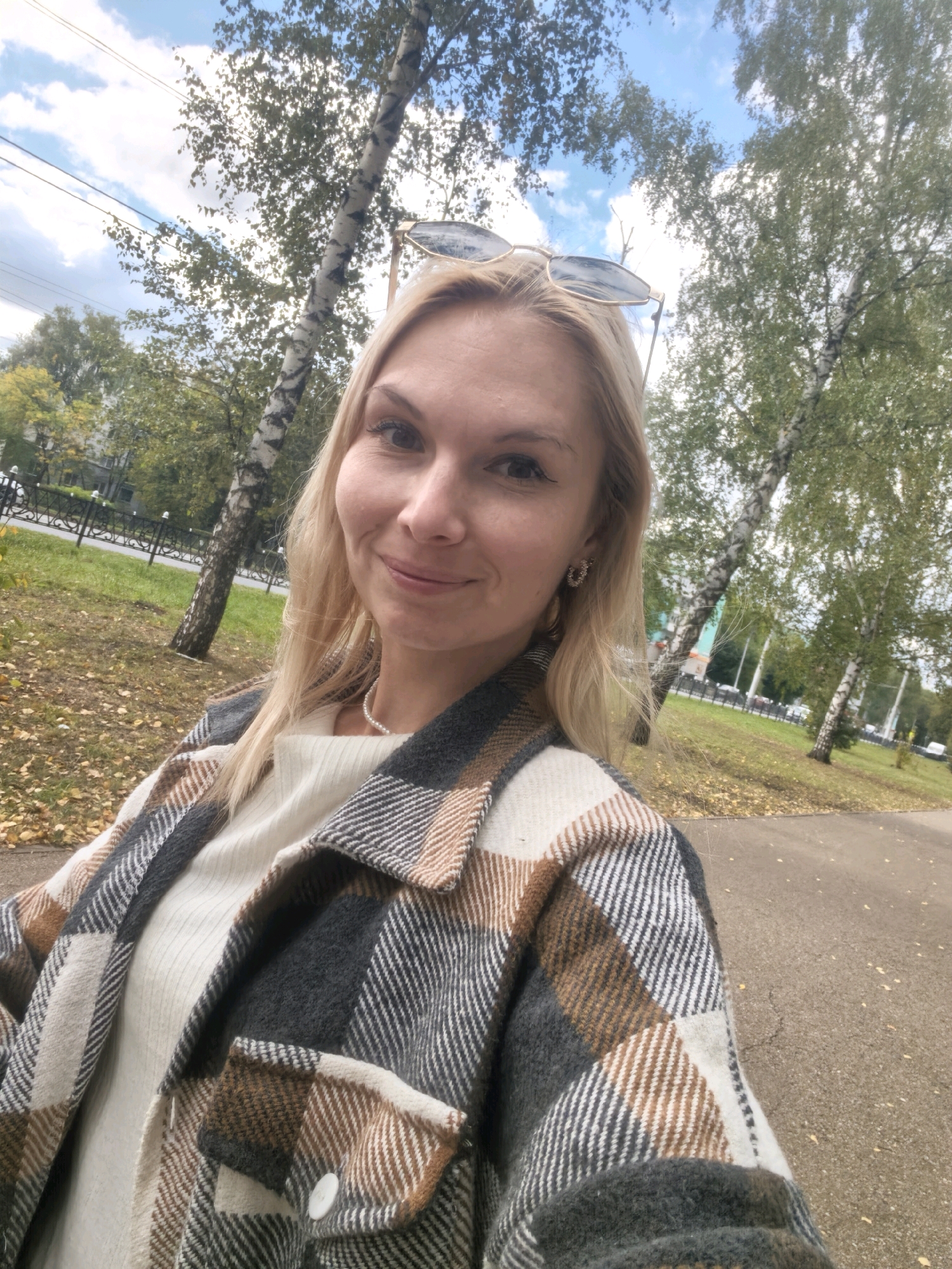 A blonde woman is taking a selfie outdoors in Sterlitamak, Russia. She is wearing a plaid jacket over a cream-colored sweater and a pearl necklace. Sunglasses are perched on her head. The background shows trees, some with autumn leaves, and a cloudy blue sky. A paved walkway and a grassy area with fallen leaves are visible in the foreground. The setting appears to be a park or a tree-lined street.