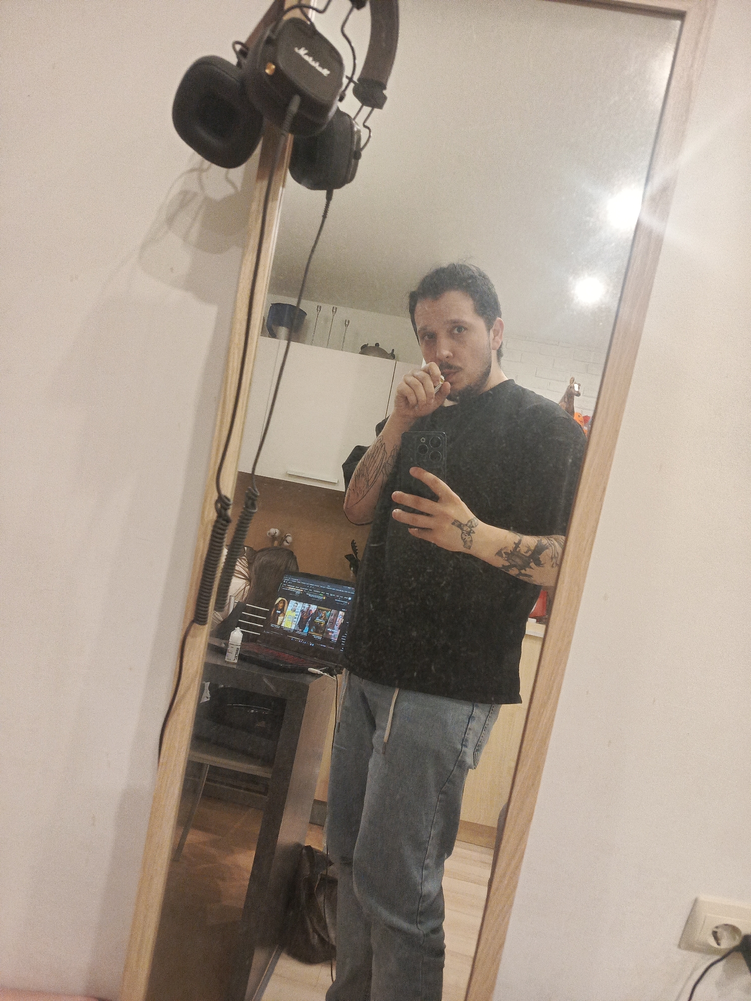 A person is standing in front of a mirror, holding a cell phone in their right hand and what appears to be a toothpick in their mouth with their left hand. They are wearing a black t-shirt and light blue jeans with a drawstring. Tattoos are visible on their forearms and hands. Reflected in the mirror is a partial view of a room with a laptop on a table. The laptop screen displays images of various people. Headphones hang from the top left of the mirror. A power outlet is visible on the bottom right.
The location is inferred to be in Penza, Russia based on the prompt.