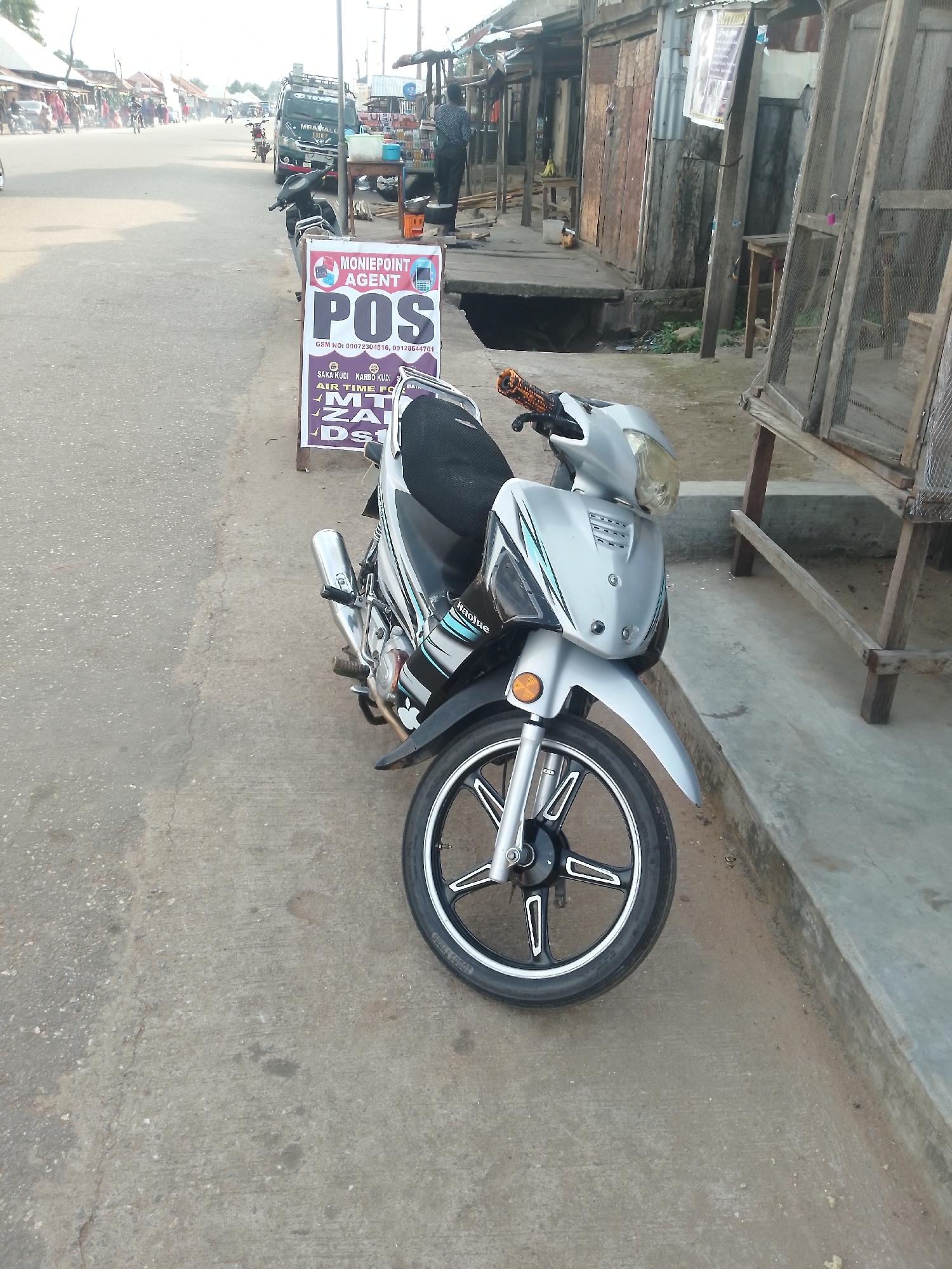 In Serti, Nigeria, a silver motorcycle is parked on a paved street in front of a row of market stalls. A "MONIEPOINT AGENT POS" sign is displayed next to the motorcycle. People are visible walking along the street and in the background. The market stalls are constructed with wood and metal, and some display goods for sale. The scene appears to be during daytime with soft natural light.