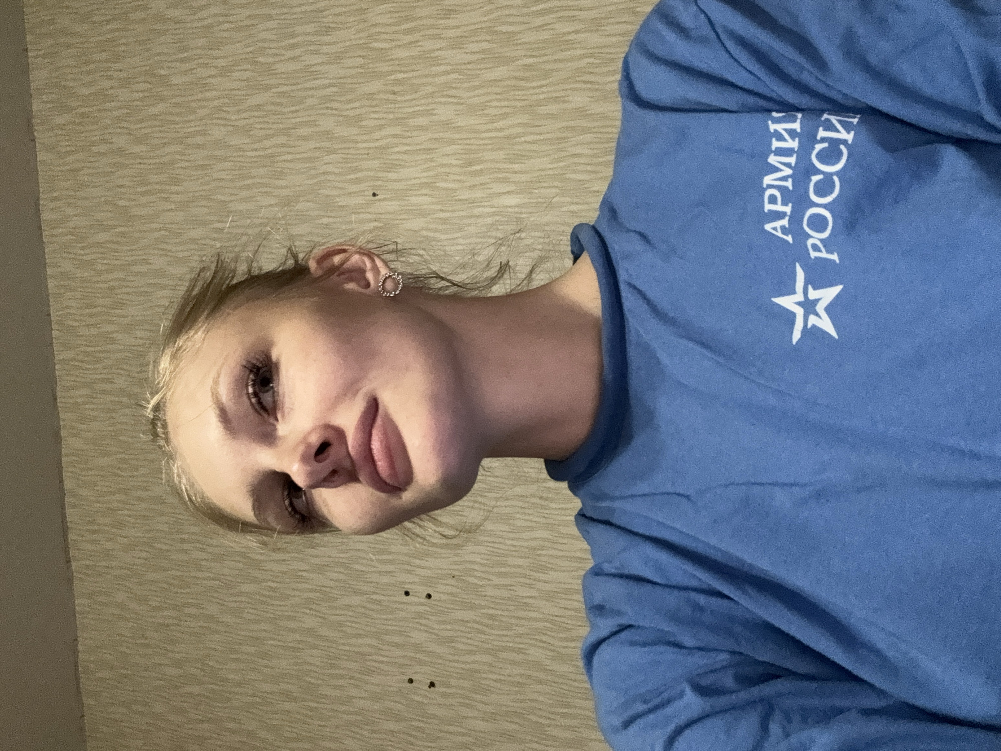 A young Caucasian woman with blonde hair tied back is looking to her left, away from the camera. She is wearing a blue t-shirt with a white emblem and text that reads "АРМИЯ РОССИИ" (Army of Russia). Her right ear is visible, adorned with a small circular earring. The background is a textured, light-brown wallpaper. There are two small dark marks on the wall behind the woman's face. The image appears to have been taken indoors. The location is specified as Kondratovo, Russia.