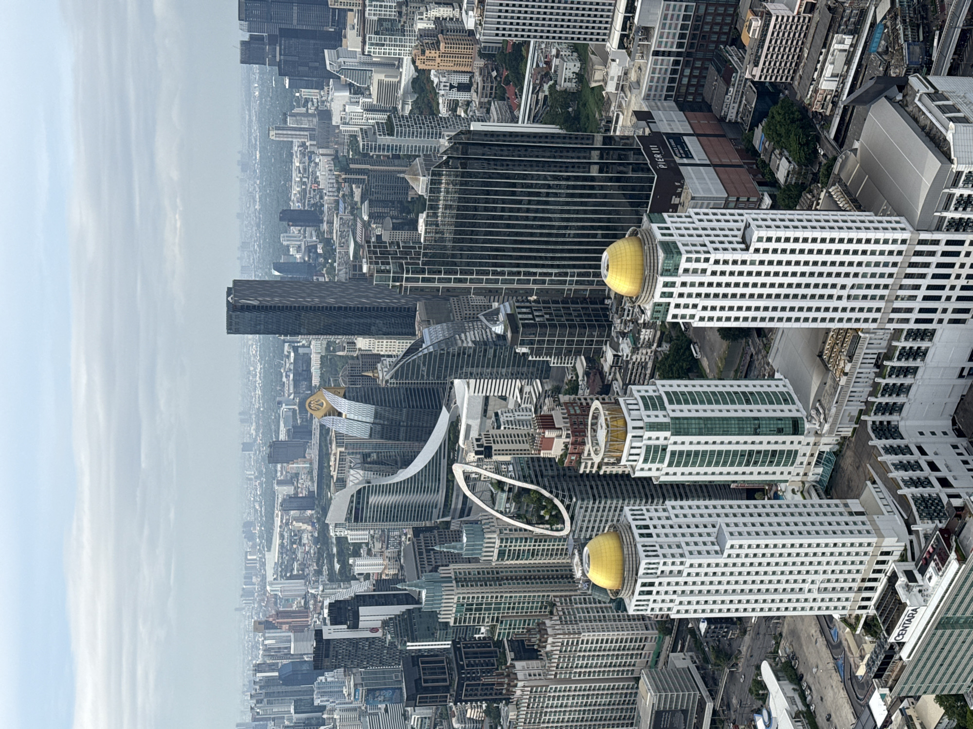 This is an aerial view of Bangkok, Thailand. Prominently featured are numerous skyscrapers and high-rise buildings. Two buildings with distinct gold domes are visible in the foreground. A distinctive architectural feature, resembling a loop, can be observed among the buildings. The environment suggests a dense urban setting with a hazy atmosphere. The image suggests a mix of modern architecture and traditional design elements. No people are visible.
