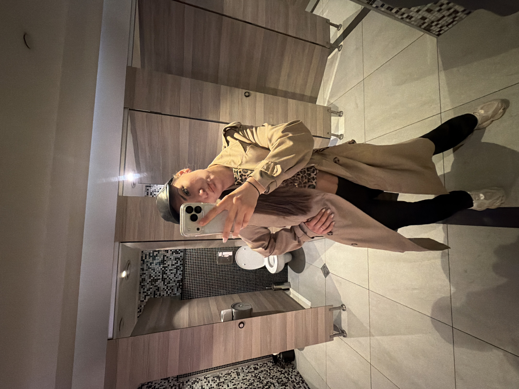 A person in a tan trench coat, black leggings, and light-colored sneakers takes a mirror selfie in a restroom. The restroom has light brown wooden stalls and white tile floors. The mirror reflects a black and white mosaic wall, a toilet, and a sink area with paper towel dispensers and other fixtures. The exact location in Sofia, Bulgaria is not discernible from the image.