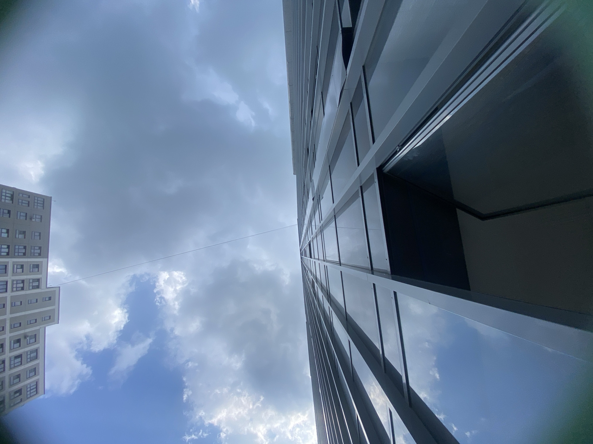 An extreme low-angle shot captures the facades of two buildings and the sky above. The building in the foreground, on the right, is a modern skyscraper with a dark glass facade featuring a grid of light-colored vertical and horizontal mullions. Its reflective surface mirrors the cloudy sky, creating a sense of depth. The building on the left, seen from a higher vantage point, appears to be a lighter-colored, multi-story residential building with numerous windows arranged in a regular pattern. A thin, dark wire stretches diagonally across the frame from the top left to the center right, connecting the two buildings or structures. The sky is partly cloudy, with patches of bright blue peeking through dramatic gray clouds. The perspective suggests the photographer is standing at the base of the skyscraper, looking upwards. The scene is set in a city environment, likely Novosibirsk, Russia, given the architectural style of the buildings.