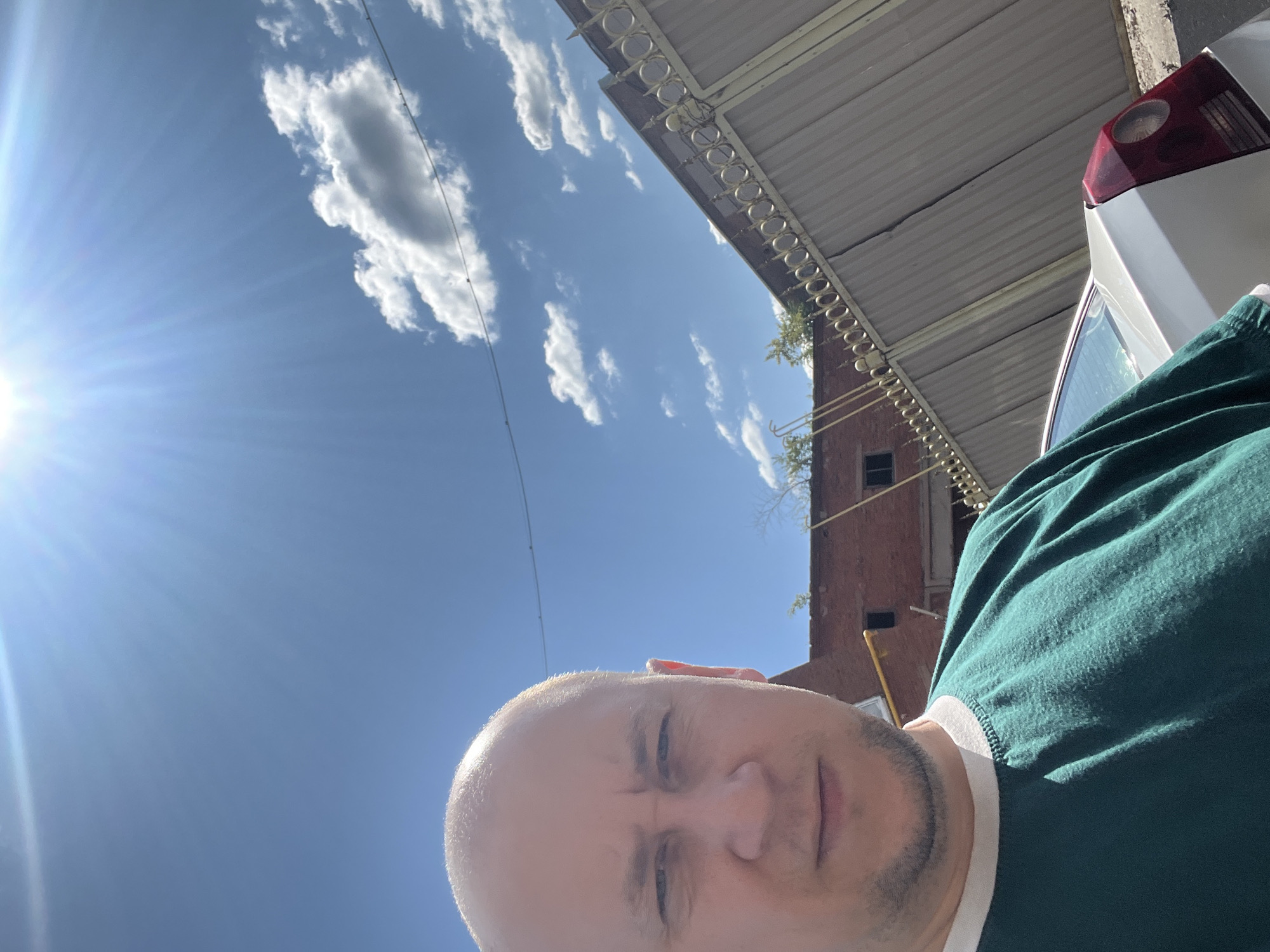 In Omsk, Russia, a man with a shaved head, wearing a teal t-shirt with a white collar, takes a selfie outdoors. The background features a brick building, a tall corrugated metal fence with decorative gold scrollwork, and a silver car. The sky is bright blue with scattered white clouds, and the sun is shining intensely, creating lens flare in the upper part of the image. The overall scene suggests a sunny day in an urban or industrial area.