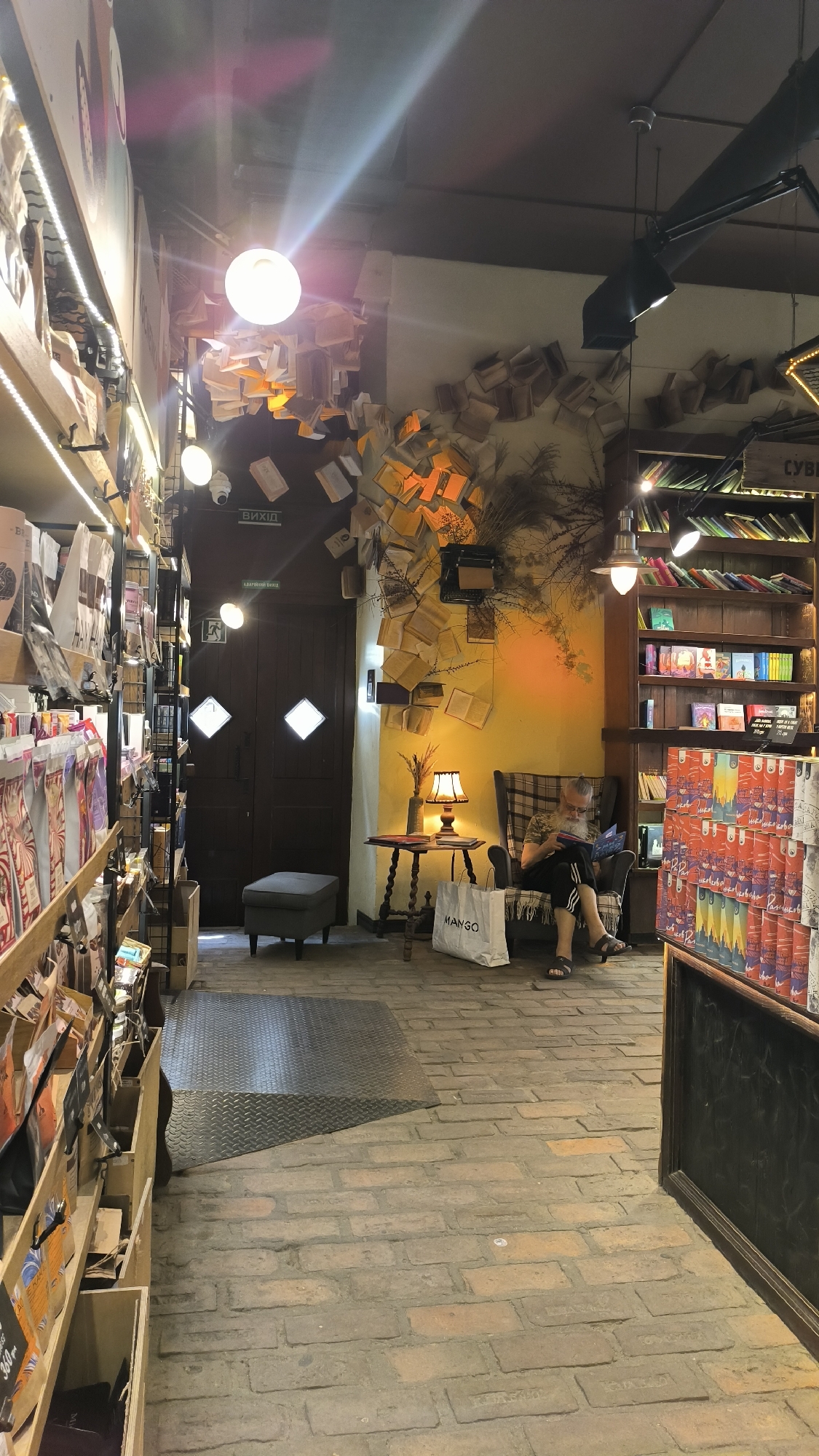 The image captures the interior of a bookstore or library with an unusual decorative theme. Books are scattered and affixed to the walls and ceiling, creating a whimsical and slightly cluttered atmosphere. The space is lit by hanging light fixtures and a lamp on a small table. A person with a long beard is seated in a plaid armchair, reading a book. To the left, shelves are stocked with various items, possibly books or merchandise. A double door is visible in the background, and a sign above it indicates "ВИХІД" (EXIT). The floor is paved with bricks, and a metal ramp is present. The overall ambiance suggests a cozy and eclectic setting.