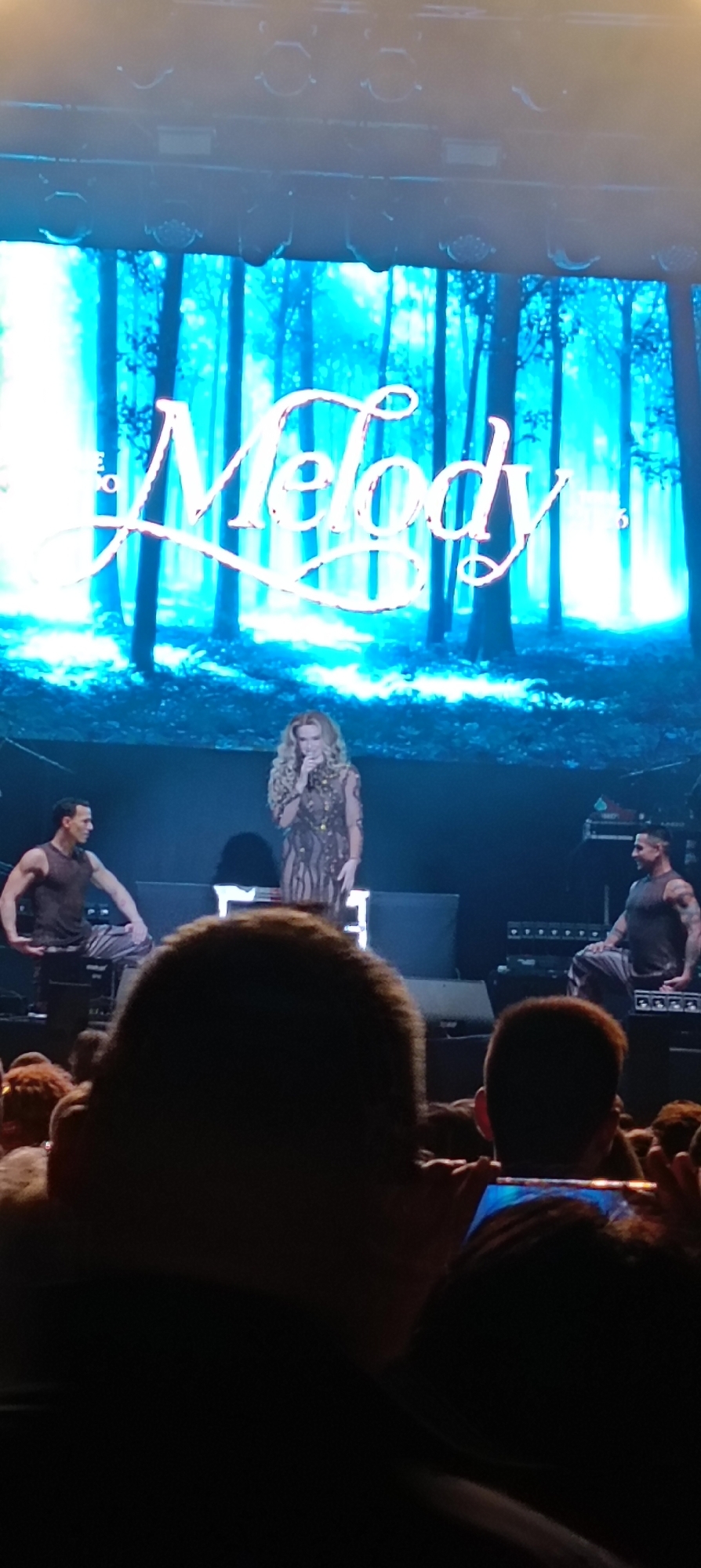 The photograph captures a live performance, likely a concert, featuring a female vocalist standing center stage. She wears a textured, possibly embellished gown and holds a microphone. Behind her, a large screen displays a stylized image of a forest with the word "Melody" superimposed in a cursive font. Two male dancers, one on each side of the stage, are dressed in tank tops and appear to be seated or posing on equipment cases. The foreground shows the heads of audience members, some of whom are holding up phones to record or photograph the performance. The lighting appears to be theatrical, with spotlights illuminating the stage and the forest screen. Based on the location of Cerdanyola Del Vallès, Spain, it might be a performance in a local venue in the town, possibly a concert or a live show.