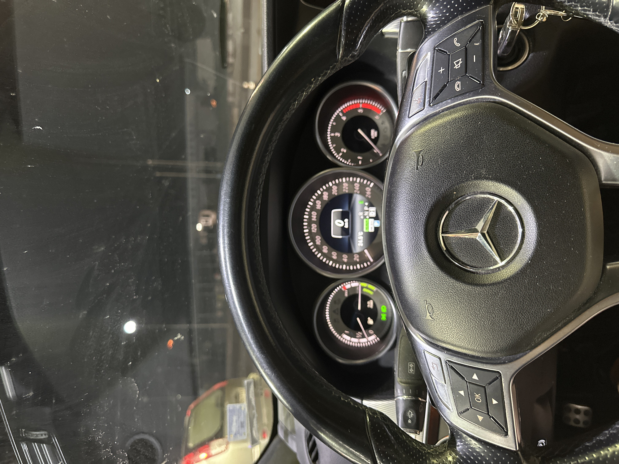 The image captures the interior perspective of a Mercedes-Benz vehicle, looking forward through a windshield. The scene is set at night or in a dimly lit indoor environment, specifically in the city of Krepost, Bulgaria.
The foreground is dominated by a black leather steering wheel featuring perforated sections on the sides and a silver metallic trim. A three-pointed star Mercedes-Benz logo is centered on the airbag cover. The left spoke incorporates a control cluster with a central "OK" button surrounded by directional arrow keys, and an additional "back" arrow button above. The right spoke contains media and telephone controls, including volume adjustments, mute, call answer/end functions, and a menu button. A set of keys, including a Mercedes-Benz smart key, is visible inserted into the ignition on the right side of the steering column. Metallic pedals with rubber inserts are partially visible in the lower-left portion of the driver's footwell.
The illuminated instrument cluster, positioned behind the steering wheel, consists of three circular gauges. The leftmost gauge indicates the fuel level, which is approximately half full, with a green "ECO" or Start/Stop indicator illuminated at its base. The central digital display shows a speed of "0 km/h" and an ambient temperature of "24.5 °C". The rightmost gauge is a tachometer, indicating the engine is at idle (0 RPM), with its redline segment beginning between 4500 and 5000 RPM. A wiper control stalk is partially visible on the left side of the steering column.
Through the vehicle's windshield, which exhibits visible smudges and dust, a dark external environment is discernible, characterized by various artificial light sources. A prominent, bright, circular white light is reflected in the upper-center of the windshield. Multiple smaller, blurred orange and red lights are scattered across the dark background, consistent with other vehicle taillights or distant urban lighting. On the left side of the view, the rear section of a light-colored vehicle, possibly an SUV or hatchback, is partially visible, displaying an illuminated taillight and an unreadable license plate frame. The vehicle is stationary, with the engine active, as indicated by the illuminated instrument cluster.