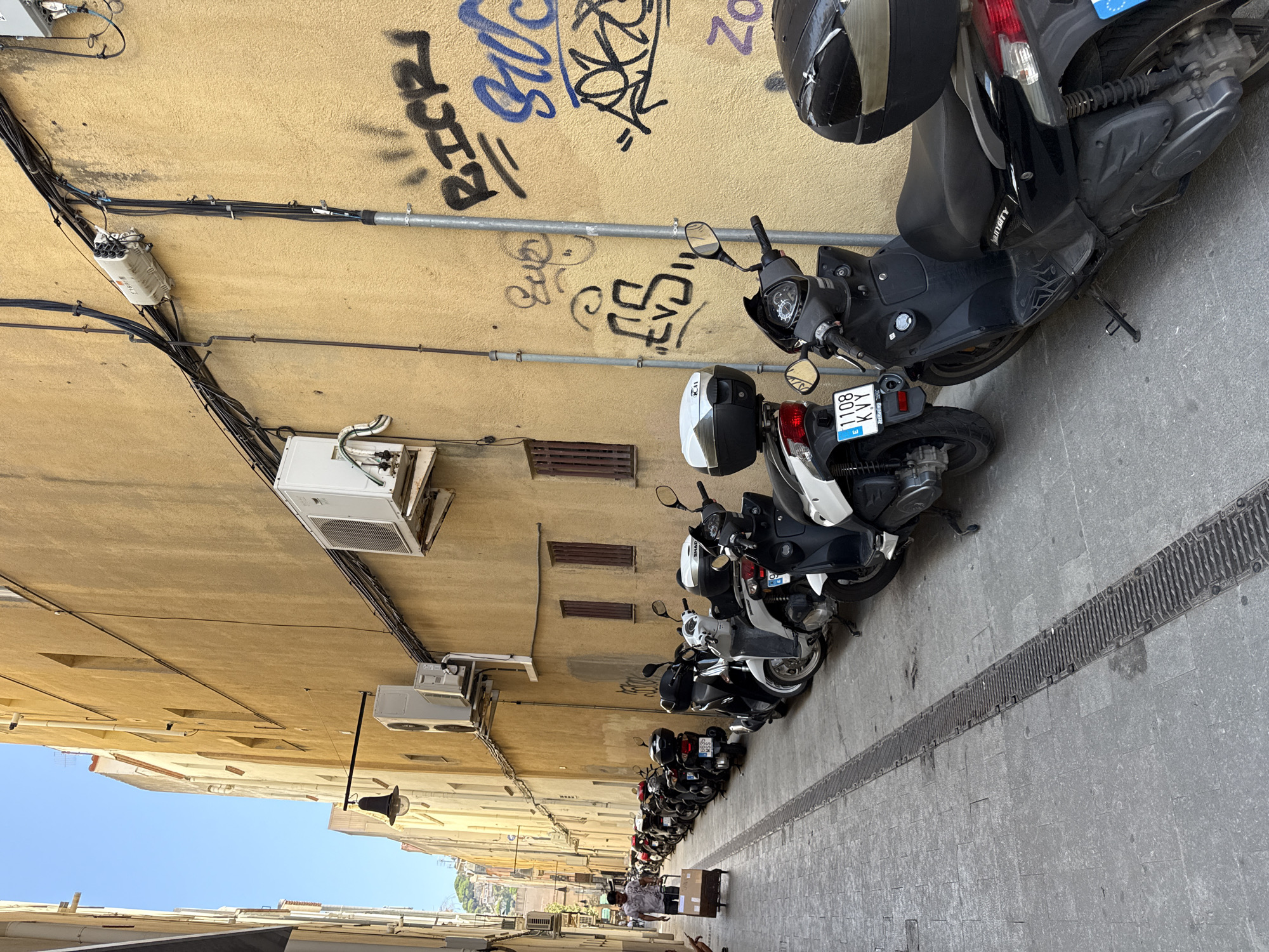 The image depicts a narrow, sun-drenched street in Blanes, Spain, lined with parked scooters and motorcycles on the right side of the frame. The building walls are painted a textured yellow, with visible graffiti tags on the right wall. Two air conditioning units are mounted on the building. A black street lamp is affixed to the wall on the left side of the street. Further down the street, a man is visible pushing a wheeled cart laden with boxes. The street pavement is made of grey stone tiles, with a metal grate running along the center, likely for drainage. The sky is a clear, bright blue.