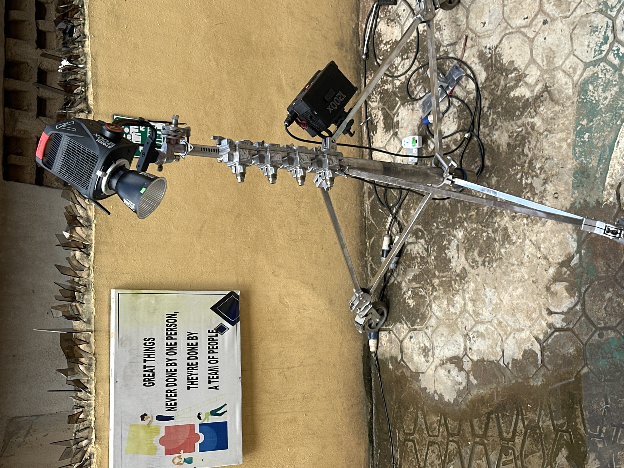 This media file captures an outdoor or semi-outdoor setup featuring professional lighting equipment, likely for a film, video, or photography shoot in Lagos, Nigeria. The primary subject is a large, black LED cinema light, identified as a "STORM 1200x" by visible text, mounted on a heavy-duty light stand. A power supply unit, also labeled "1200x," is attached to the stand. The stand is equipped with multiple clamps and rigging accessories.
The setting includes a light brown/tan wall, topped with a row of anti-climb spikes made of broken glass or similar sharp objects. Leaning against this wall is a motivational poster displaying colorful graphic elements and the text: "GREAT THINGS NEVER DONE BY ONE PERSON, THEY'RE DONE BY A TEAM OF PEOPLE." The ground is paved with hexagonal concrete tiles, appearing damp and somewhat discolored in areas, suggesting recent moisture or heavy use. Various black cables for power and equipment are loosely strewn across the tiled ground.
No people are visible in the image, but the arrangement of the equipment strongly implies a production activity is either ongoing, has recently concluded, or is set up to begin. The time of day appears to be daylight, given the general illumination, though specific weather conditions are not clear beyond the damp ground. The overall scene suggests a functional, behind-the-scenes environment of a production set.