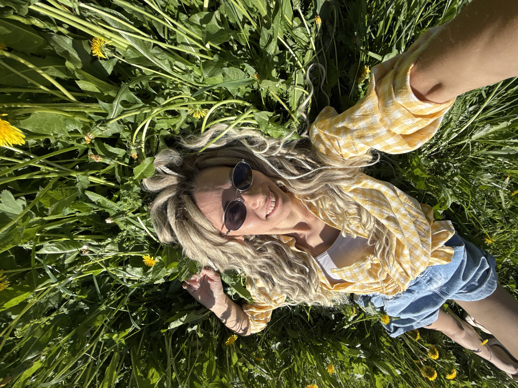 A woman lies in a field of green grass and yellow dandelions. She wears sunglasses, a yellow and white checkered shirt, and denim shorts. She is smiling and appears to be taking a selfie. Her location is Шебунино, Russia.