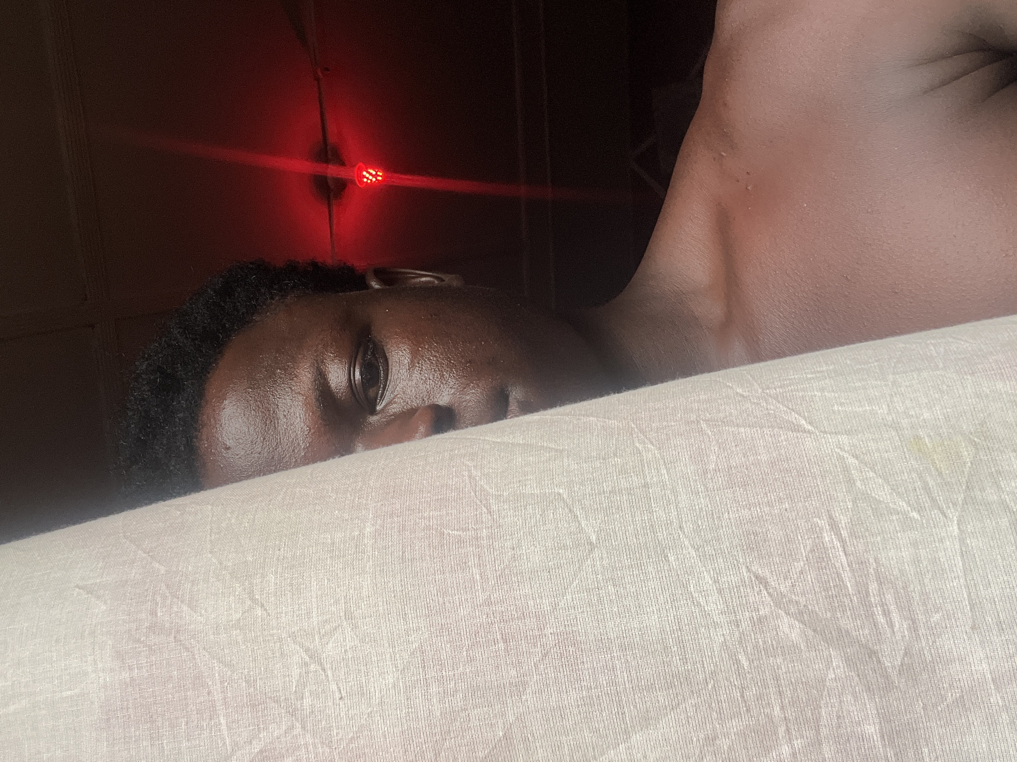 A dark-skinned person with short, dark hair is lying down on a light-colored, textured surface. The person's face is partially visible, with one eye open and facing the camera. The background is dark, with a red light visible on the wall. The person's chest and shoulder are also visible. The location is not discernible from the image. There are no apparent actions or interactions taking place. The environment is dimly lit and lacks recognizable details.